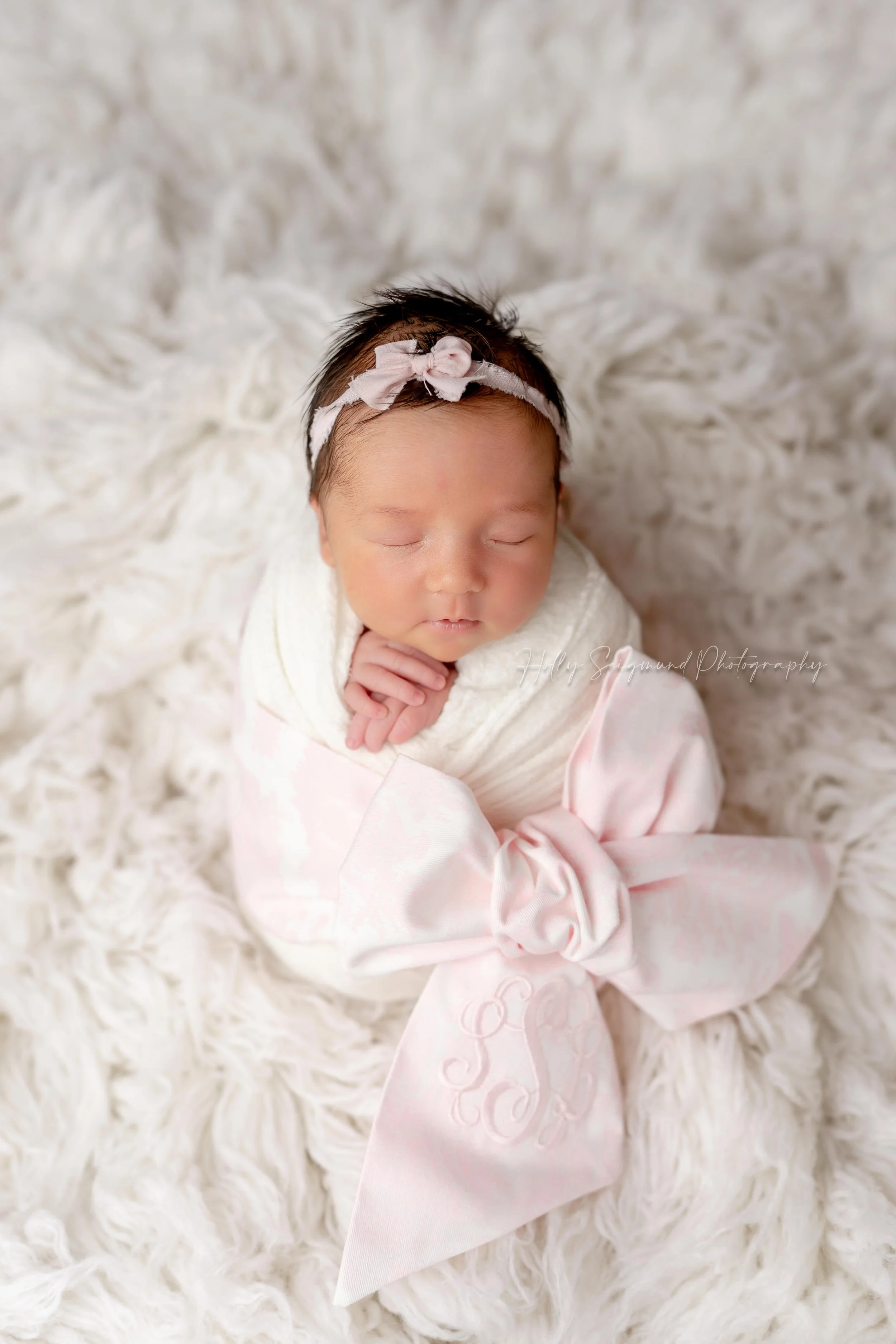 Newborn photography serving Elgin, Texas featuring a baby girl wrapped in white with a monogrammed pink bow, on a fluffy white backdrop and a small pink bow