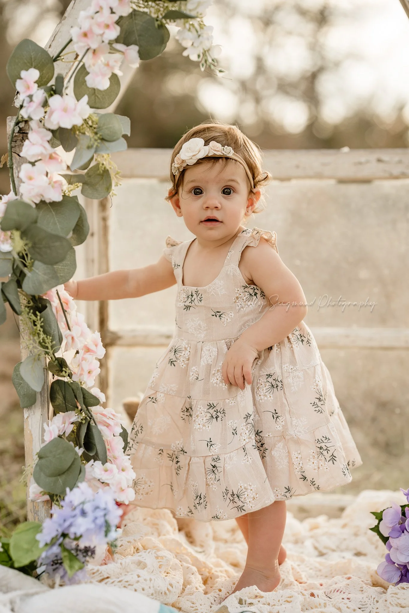 One year old girl in vintage dress with floral headband in a mini greenhouse with flowers in Lexington, Texas
