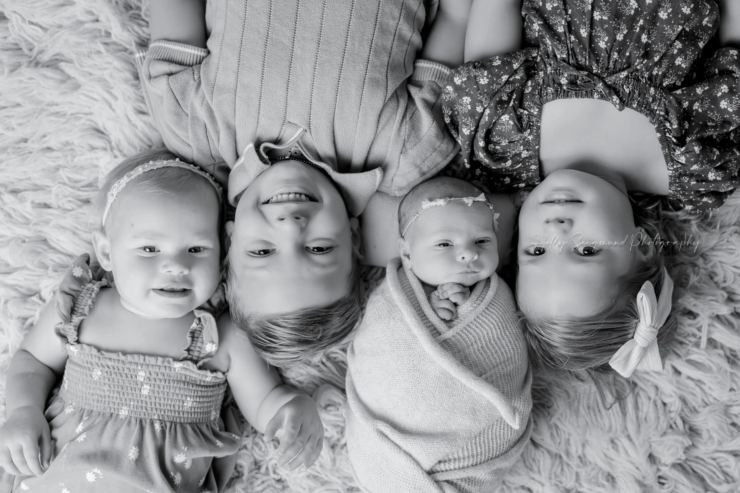 4 siblings in a black and white image, all laying on their backs on a fluffy rug, looking up at the camera during a in-home newborn photography session in Dripping Springs, Texas