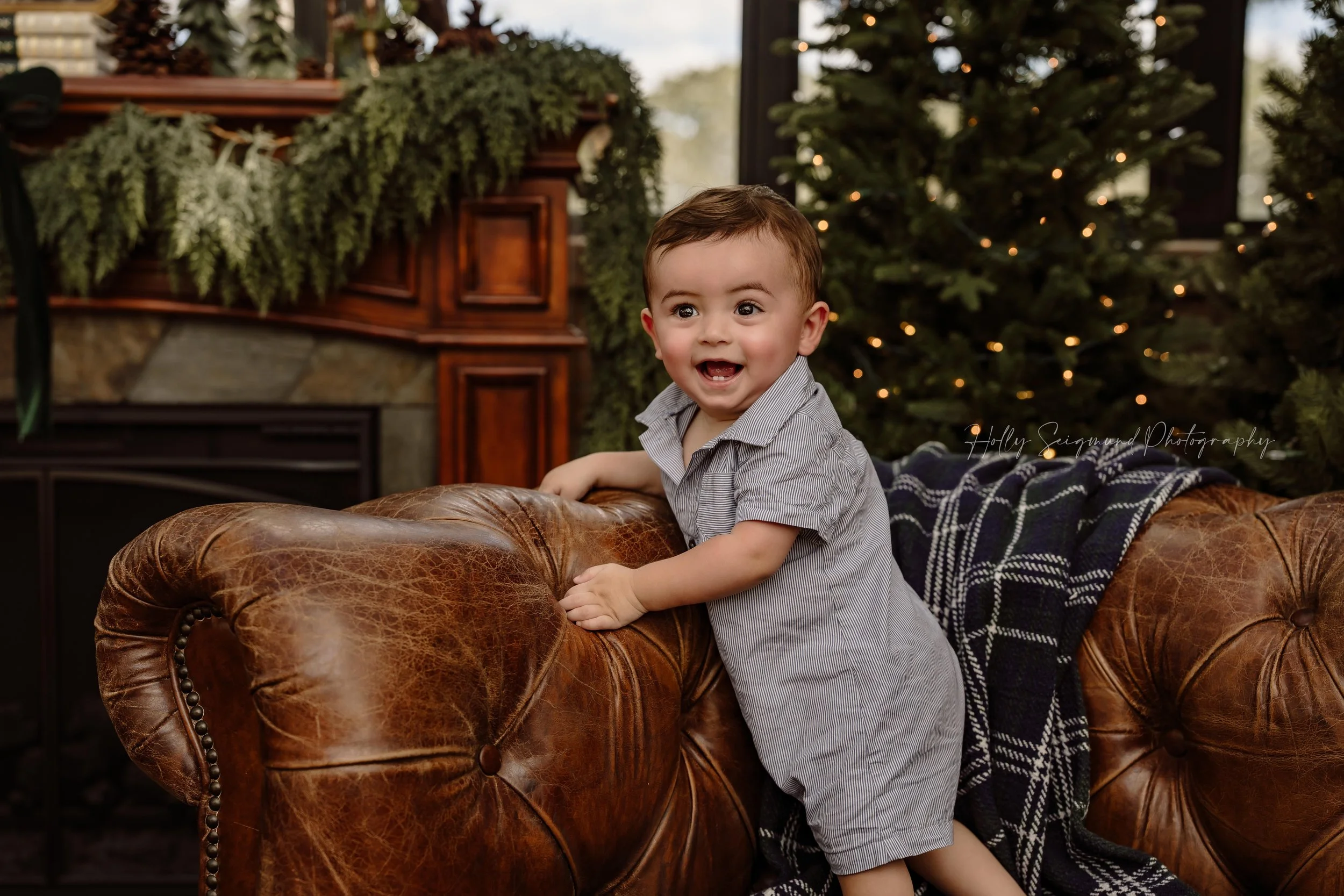 Smiling baby boy in a light blue romper standing against a brown leather sofa, with softly glowing Christmas trees behind him during a kids' Christmas mini session with Holly Seigmund Photography in Blue, Texas.