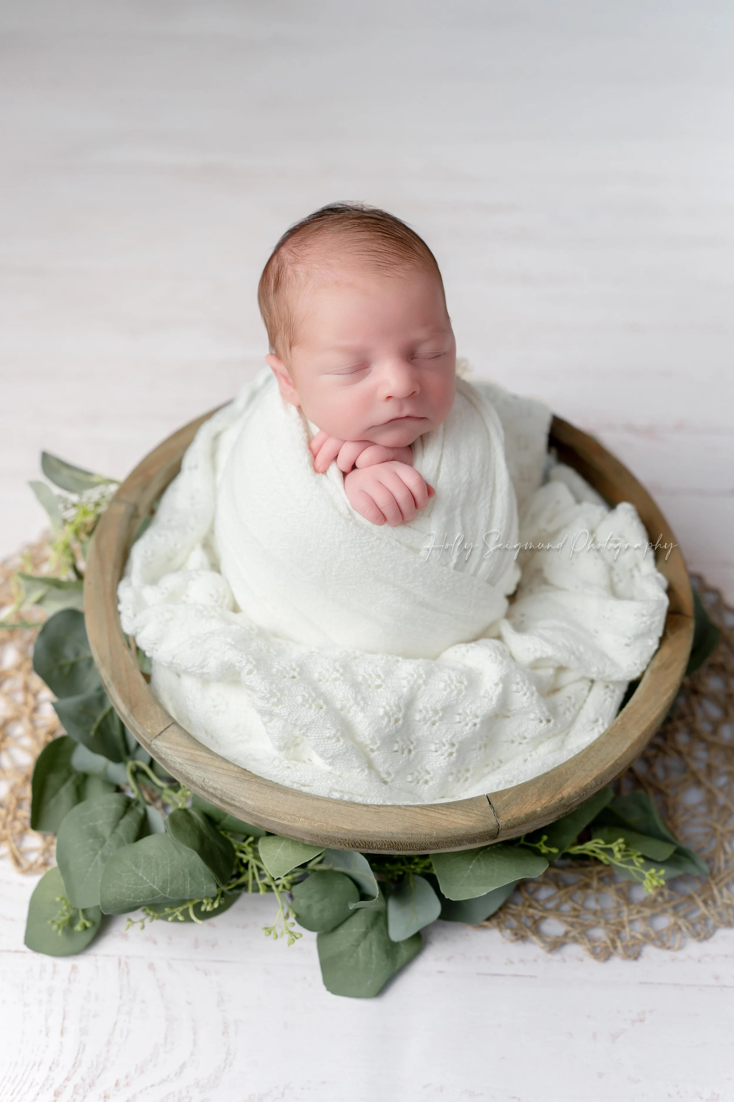 Baby newborn boy wrapped in a white wrap, posed in a wooden bowl with greenery during his in-studio newborn session in Elgin, Texas