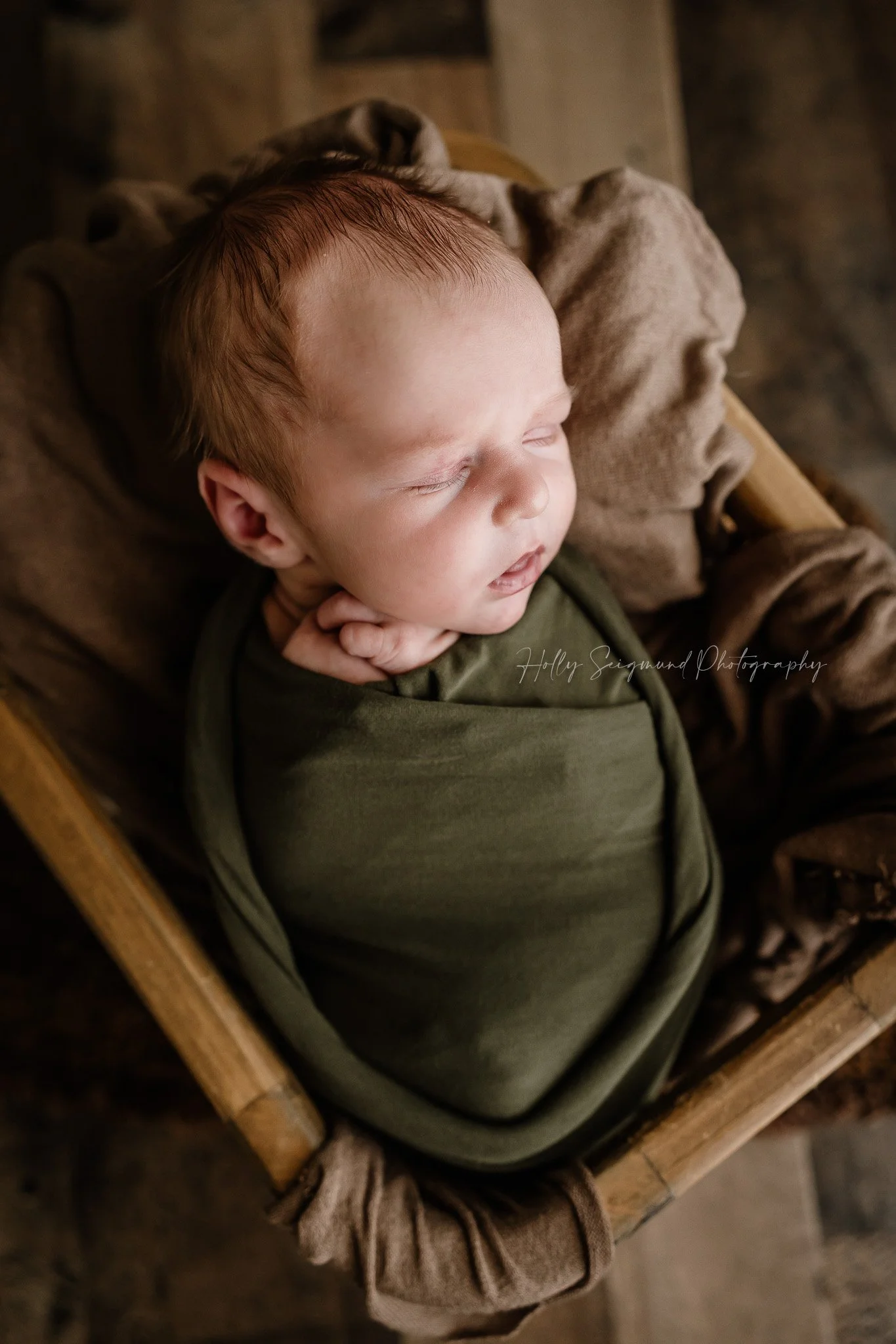Newborn baby bow wrapped in green, in a wood basket on a wood floor in Lee County, Texas