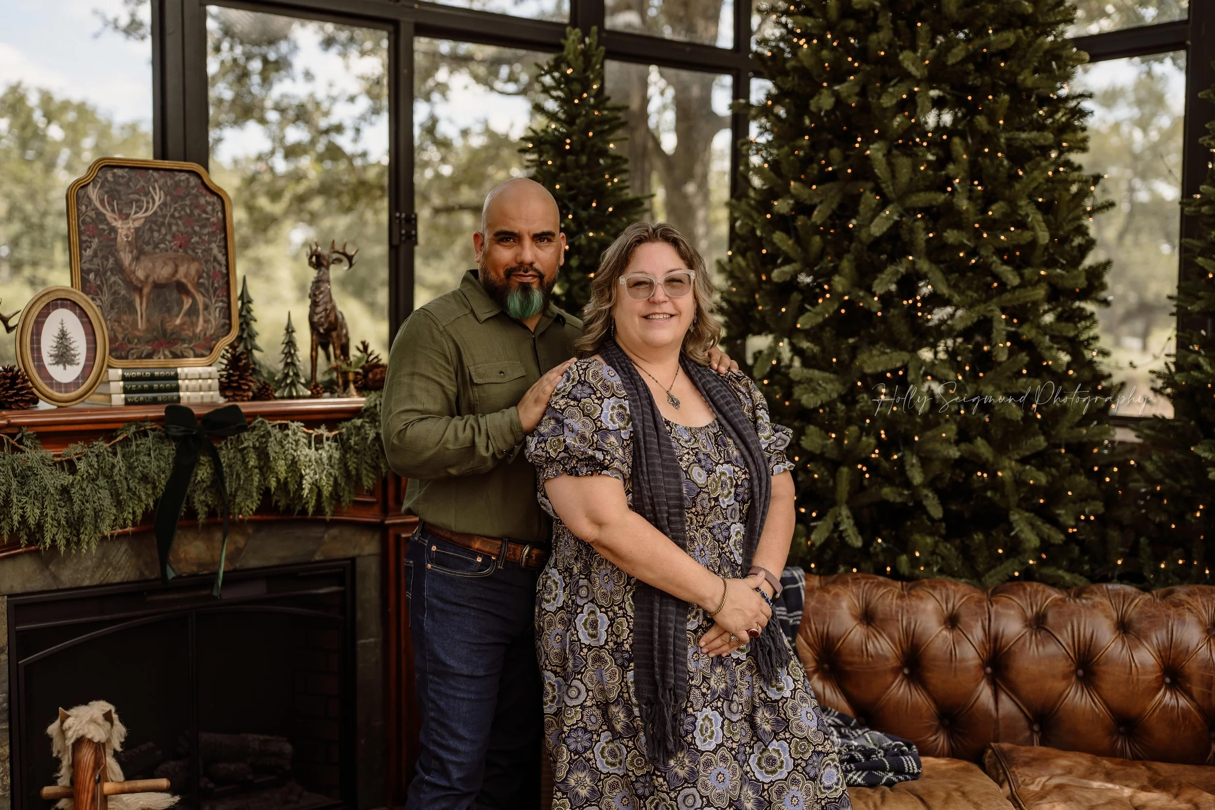 Couple posing together beside a brown leather sofa and Christmas trees with twinkling lights during a holiday mini session with Holly Seigmund Photography in Blue, Texas.