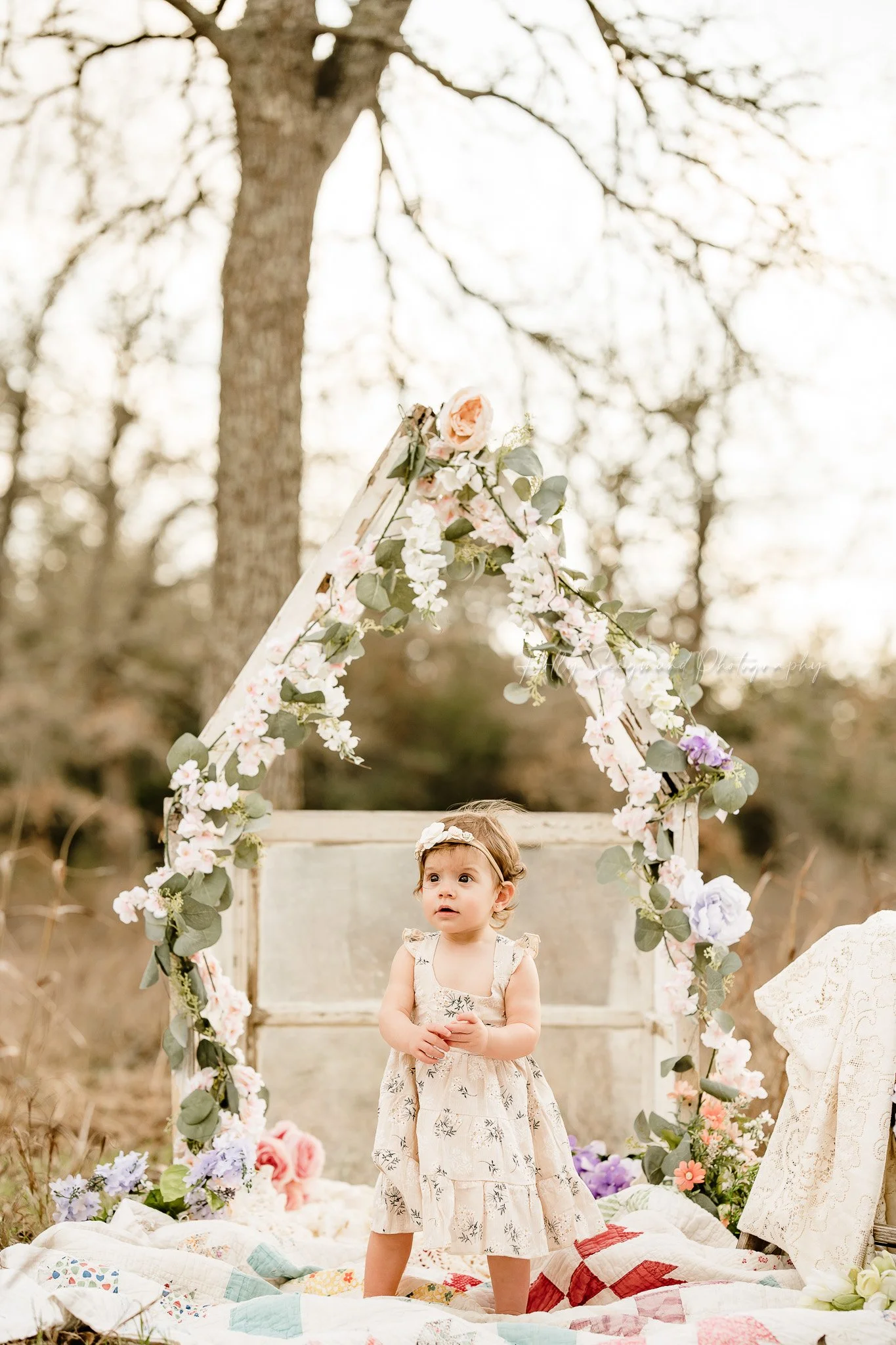First birthday girl on a quilt in front of a mini greenhouse with florals
