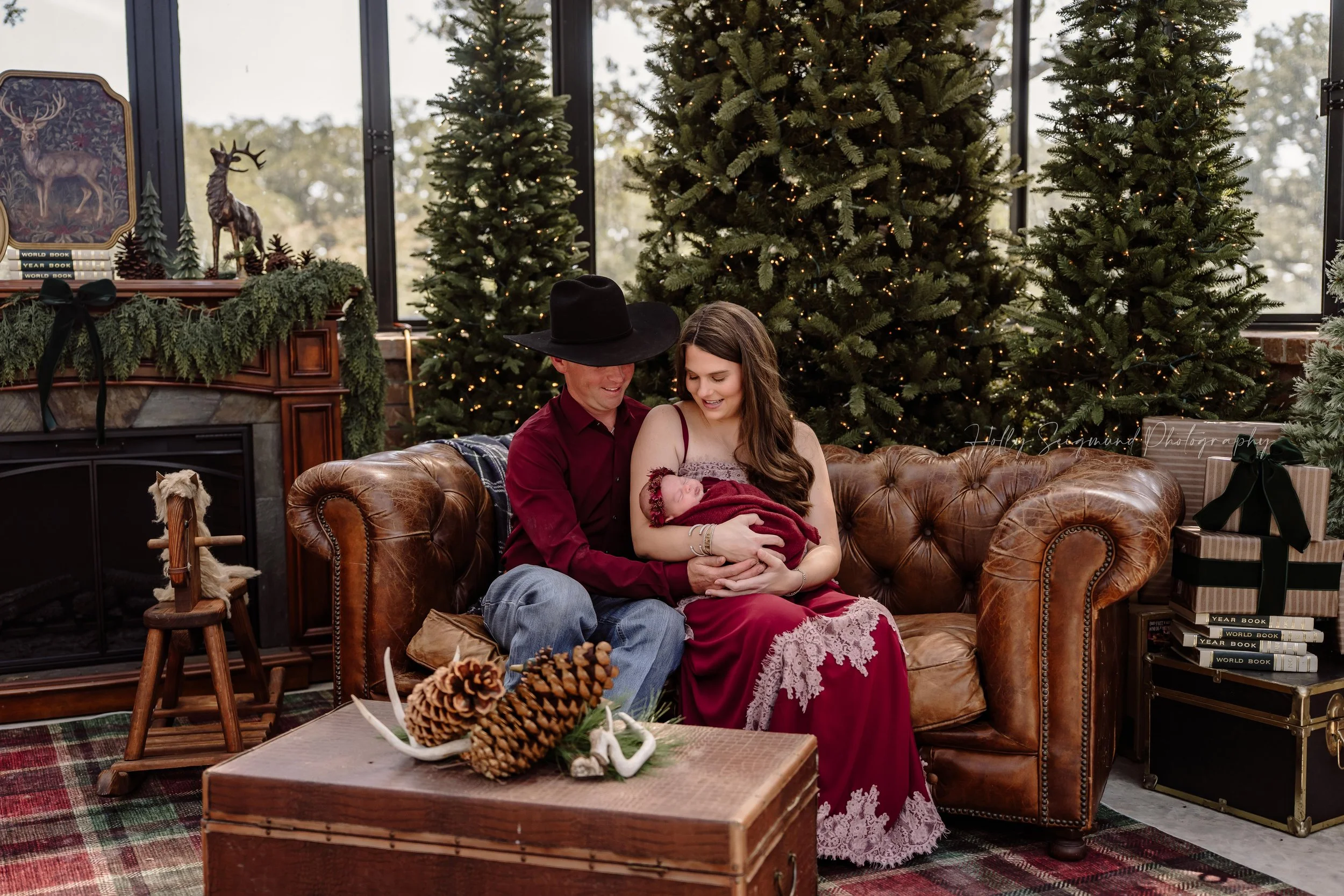 Parents smiling at their newborn baby wrapped in burgundy while sitting on a brown leather sofa, surrounded by Christmas trees during a family Christmas mini session with Holly Seigmund Photography in Blue, Texas.