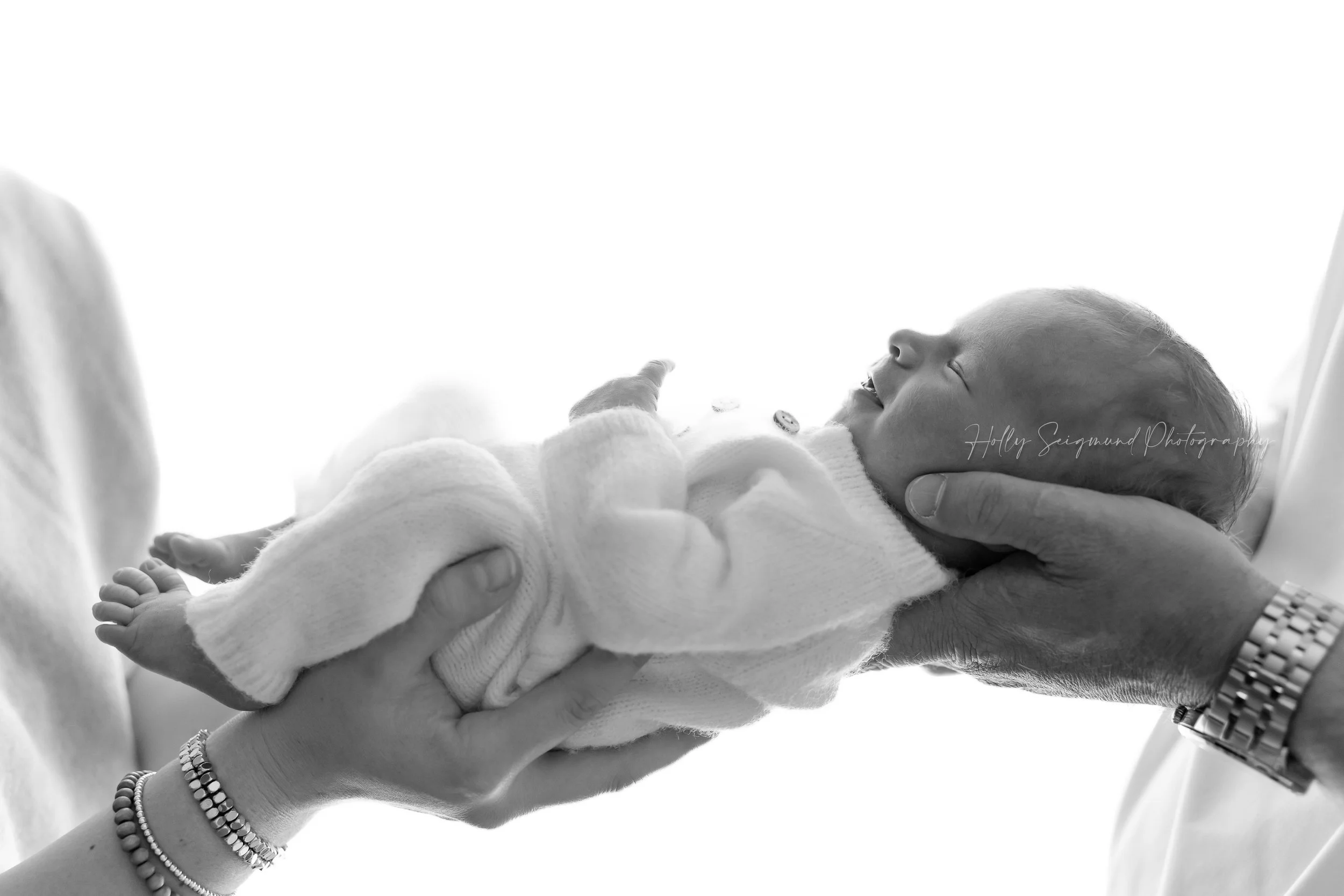 Black and white photo of a newborn baby girl in a white knit outfit, held in mom and dad's hands during a family newborn photography session serving Bastrop, Texas