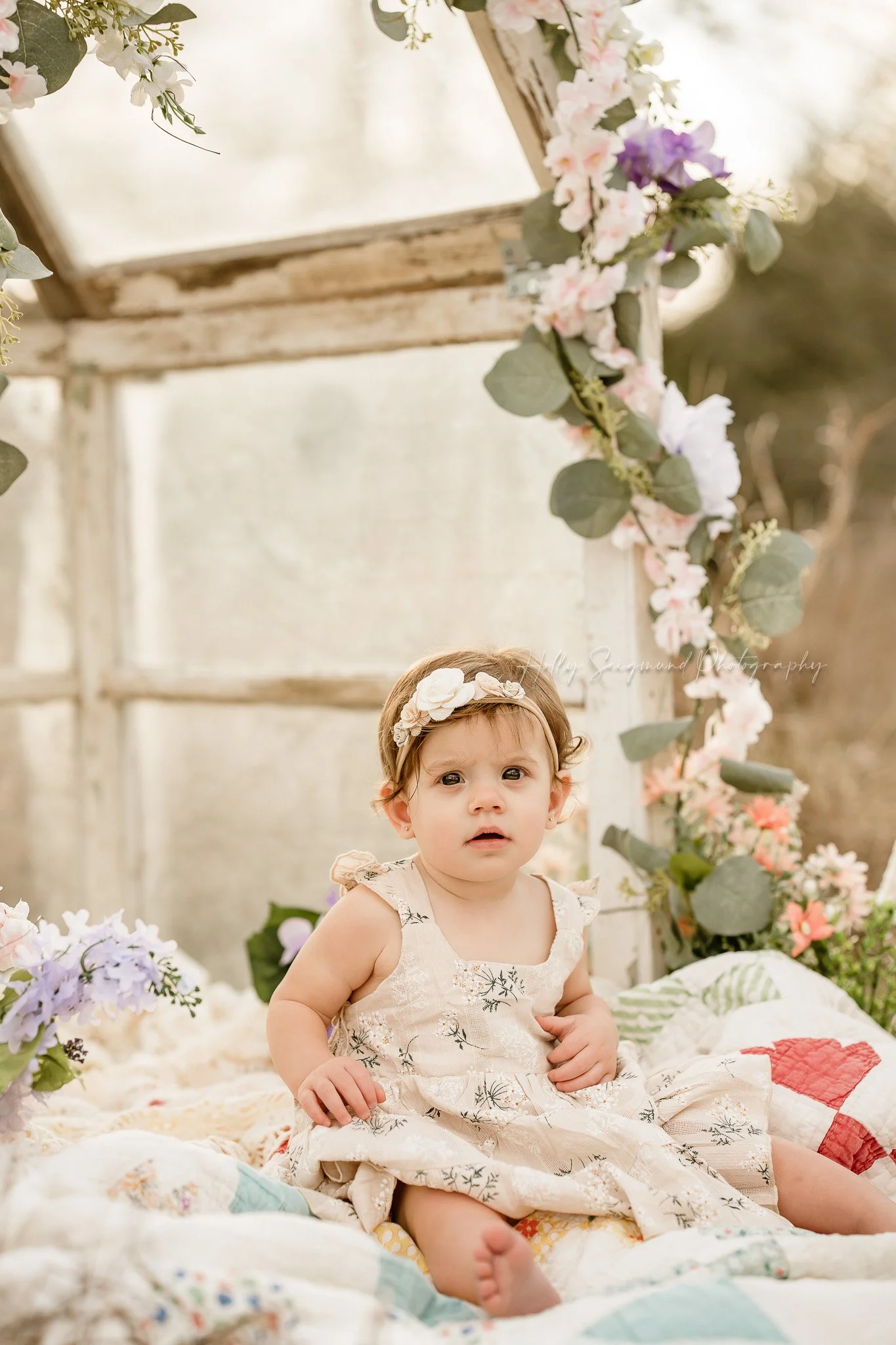 One year old girl on a vintage quilt in front of a mini greenhouse covered in florals in Lee County Texas