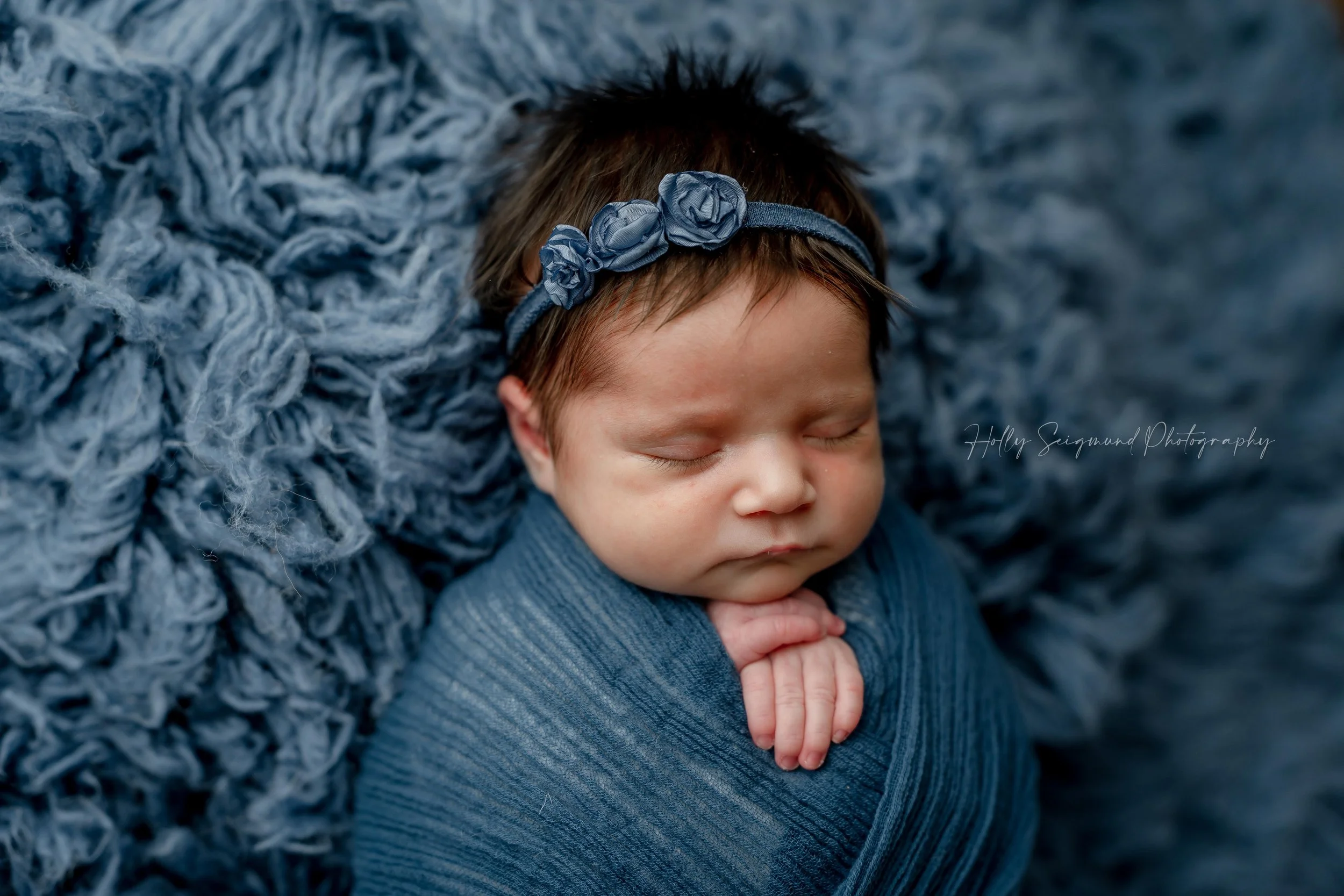 Baby girl with lots of hair on a true blue fluffy backdrop, blue wrap and blue flower headband during a studio newborn session in Blue, Texas