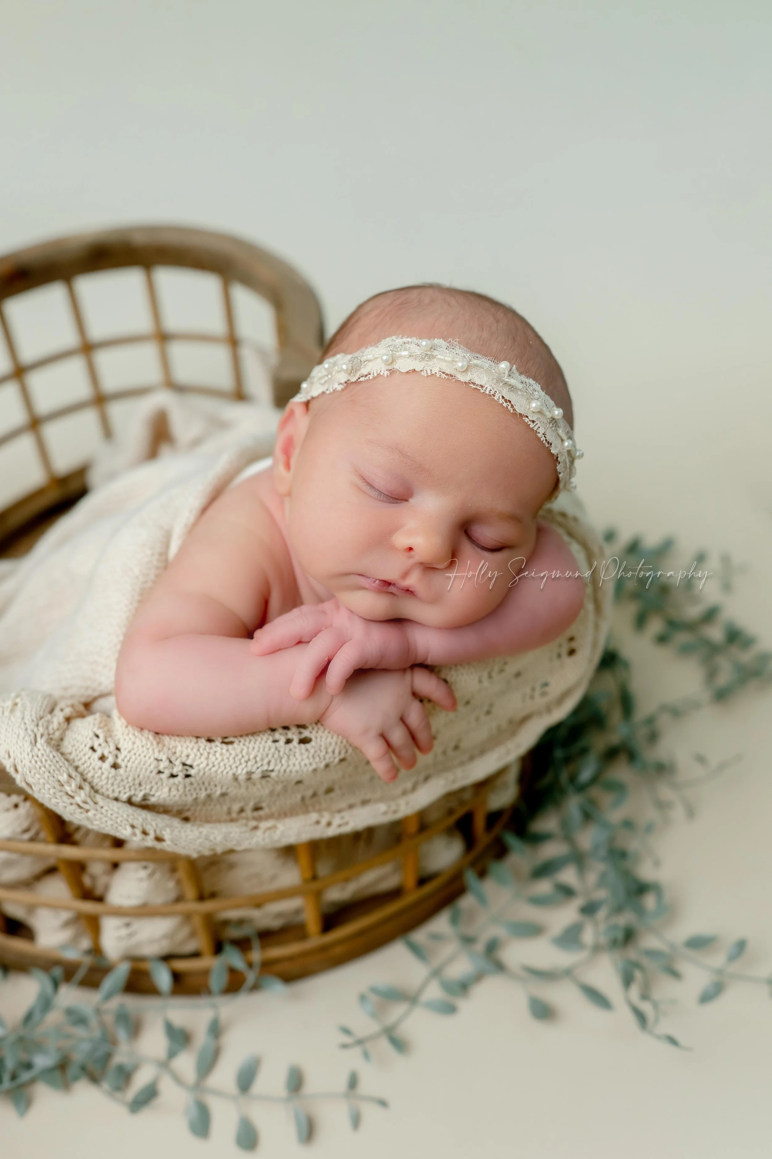 Tiny little baby girl with a cream lace headband, posed in a wooden heart crate decorated with greenery on a neutral backdrop during a in-home studio newborn photography session in Dripping Springs, Texas
