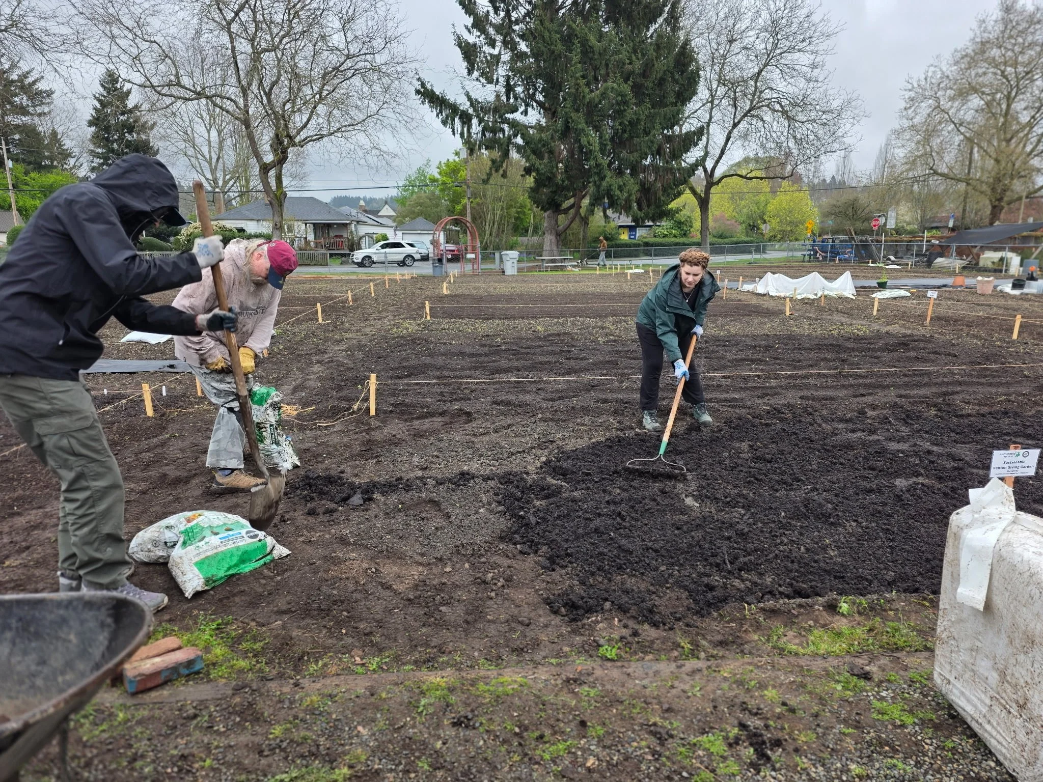 People working in a garden or farm preparing soil for planting, with trees, houses, and cloudy sky in the background.