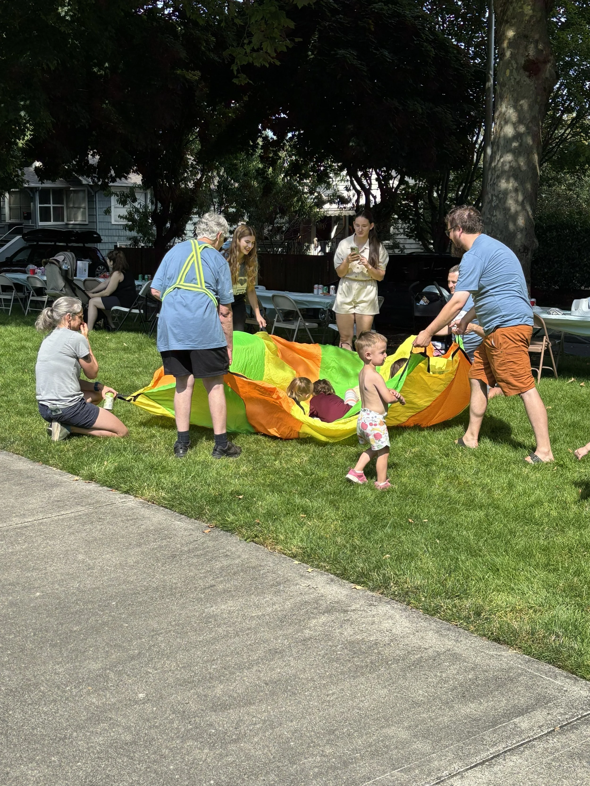 A group of people including children and adults gathered outside in a backyard, participating in a game with a colorful parachute. There are tables with food and drinks, and trees providing shade in the background.