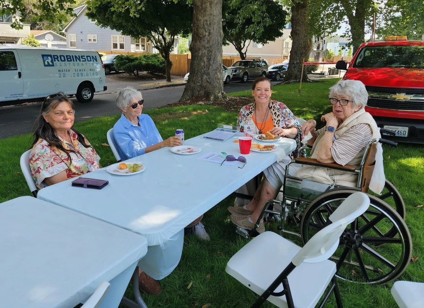 Four women sitting around a picnic table outdoors, enjoying a meal together on a sunny day, with trees and parked cars in the background.