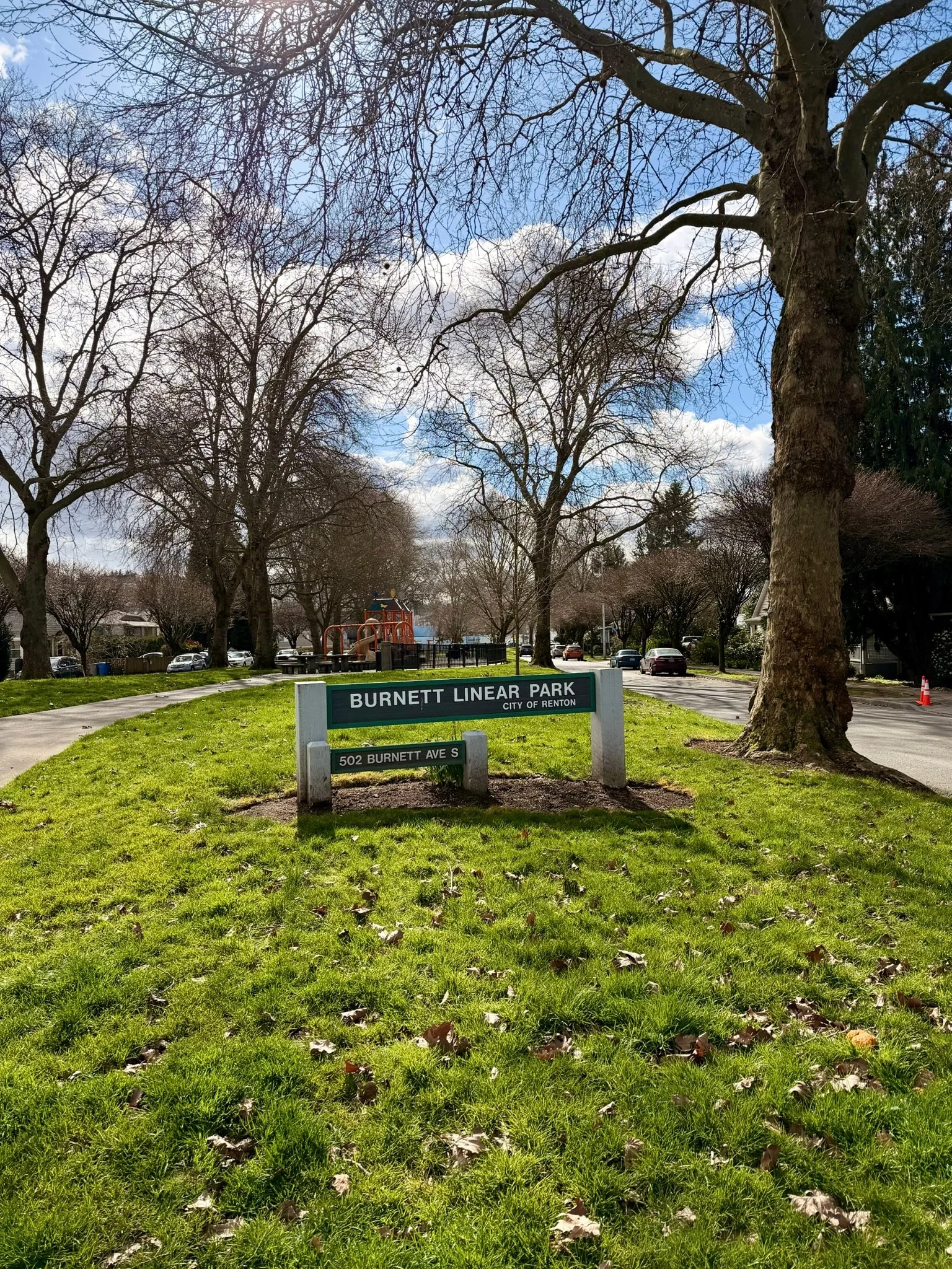 A park scene with leafless trees, a grassy area, a playground, and parked cars, with a sign that reads 'Burnett Linear Park, City of Renton' at 502 Burnett Ave S.