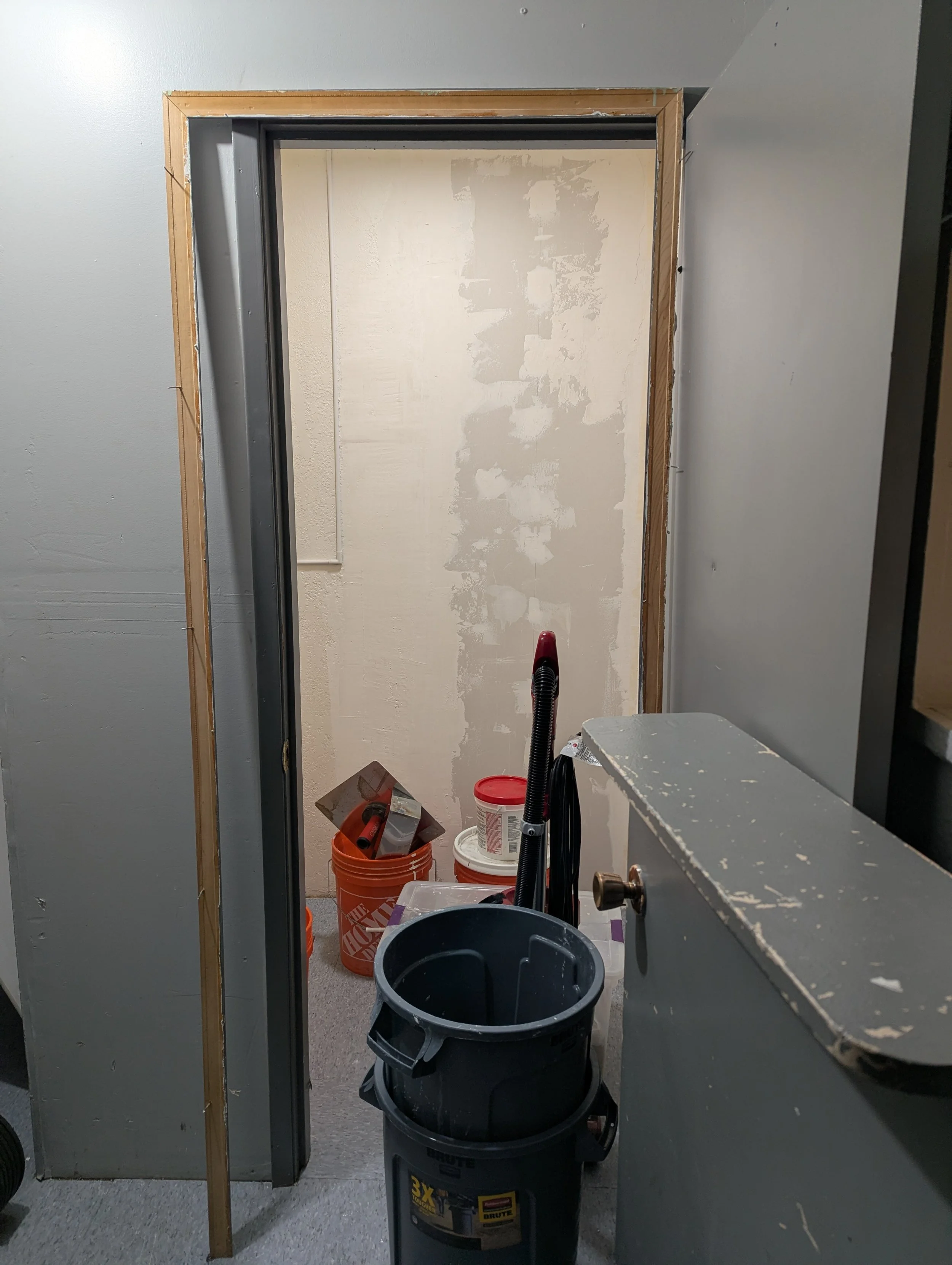 Construction site with wall in progress, buckets of paint, tools, and debris, viewed through a doorway.