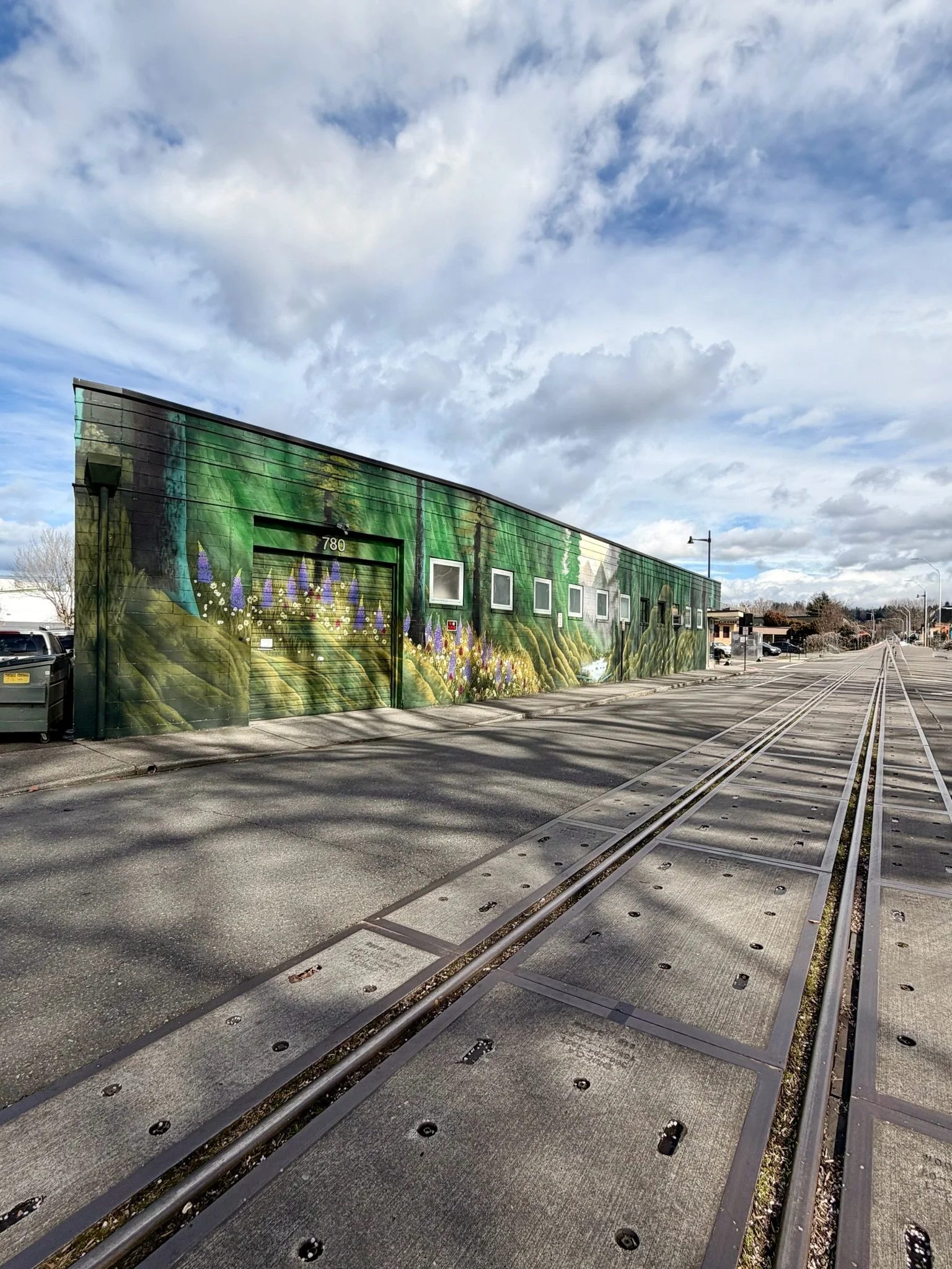 A building with a mural of a green forest and lavender flowers painted on the wall, next to a parking lot and streetcar tracks.