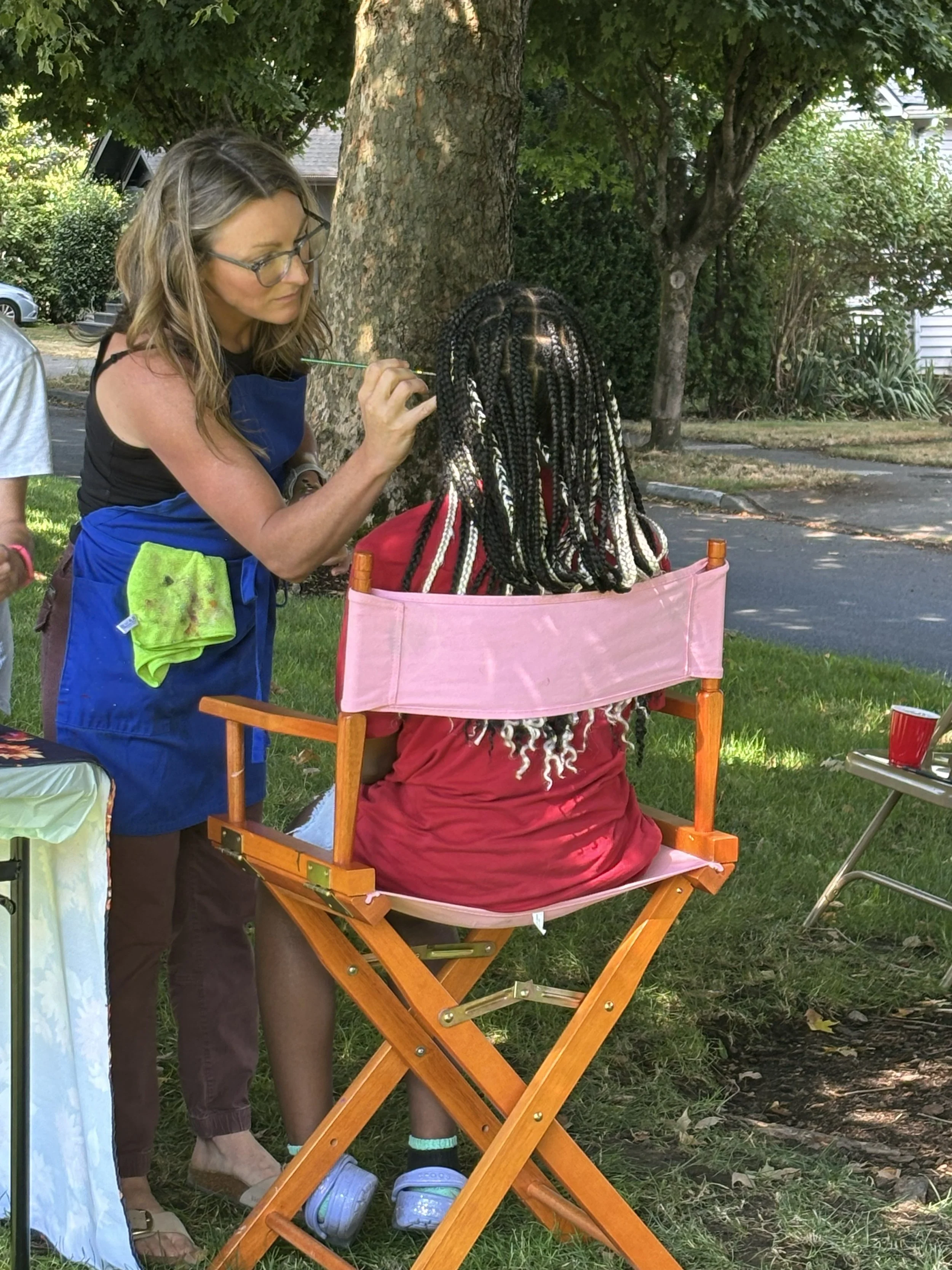 A woman with glasses applying makeup or face paint to a person's face outdoors near a large tree, with other trees and a house in the background, and a pink folding chair.