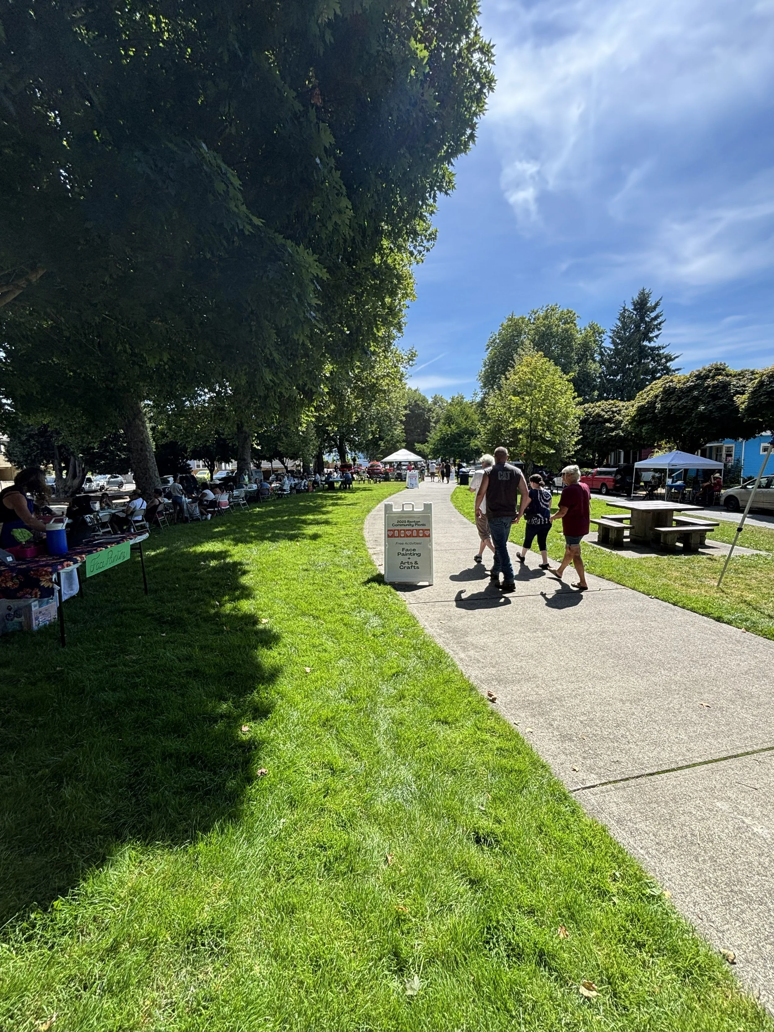 A park festival scene with tents, trees, and people walking, sitting, and enjoying an outdoor community event on a sunny day.
