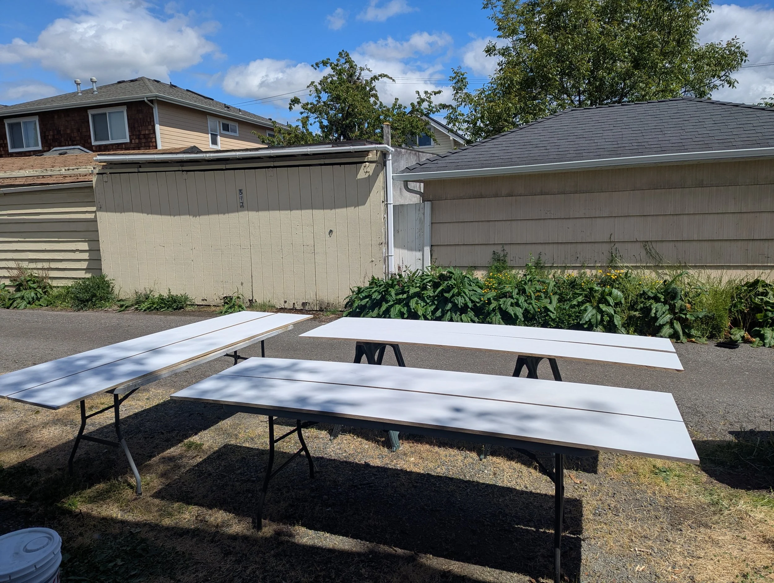 Outdoor scene with white folding tables set up on a grass and gravel surface, with houses and trees in the background under a partly cloudy sky.