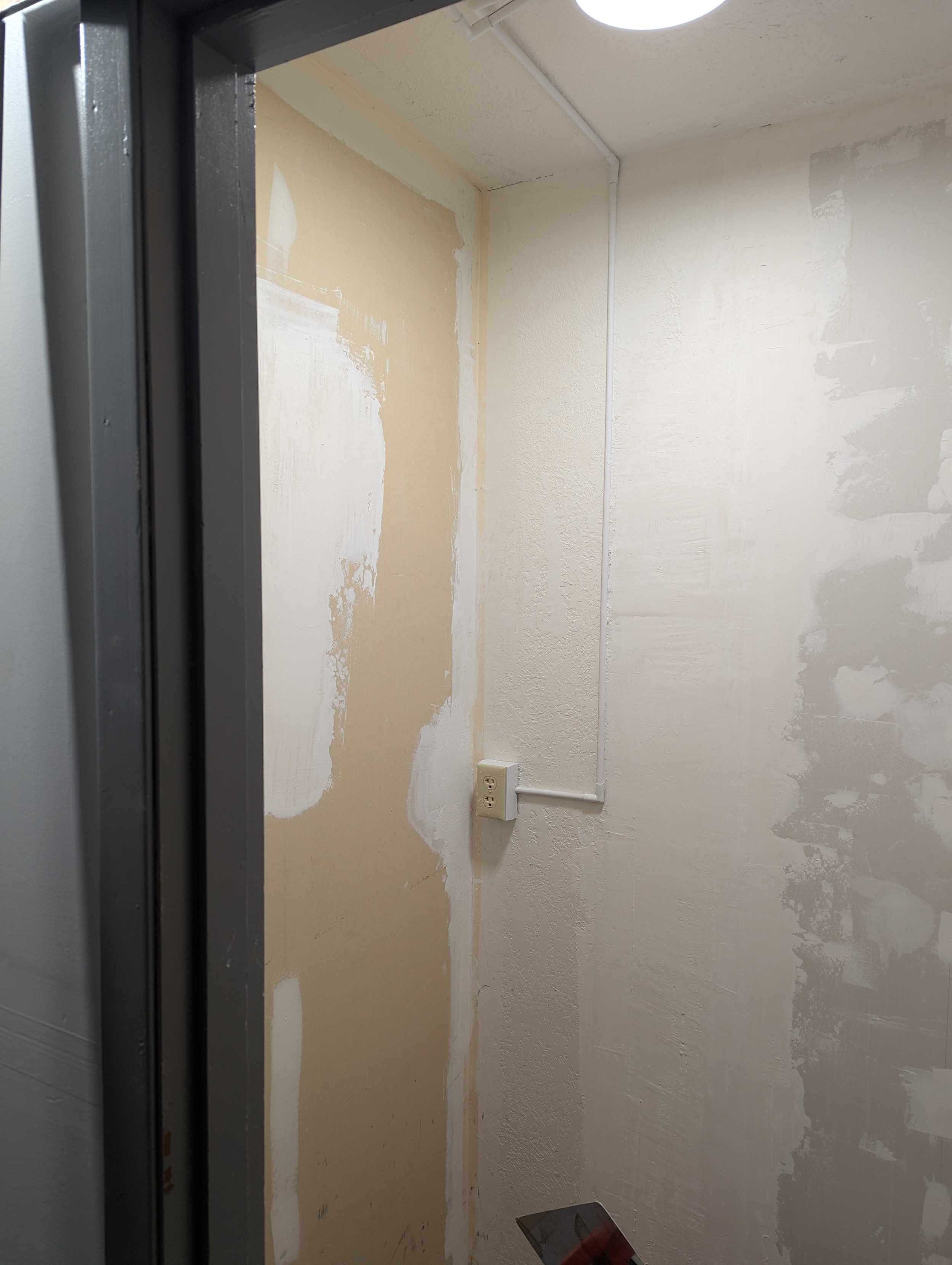 An empty room with textured walls painted in a light cream color, with some areas appearing to be patched or under renovation. There is a black electrical outlet on the lower part of the wall, and a white cable runs vertically along the wall. A ceili