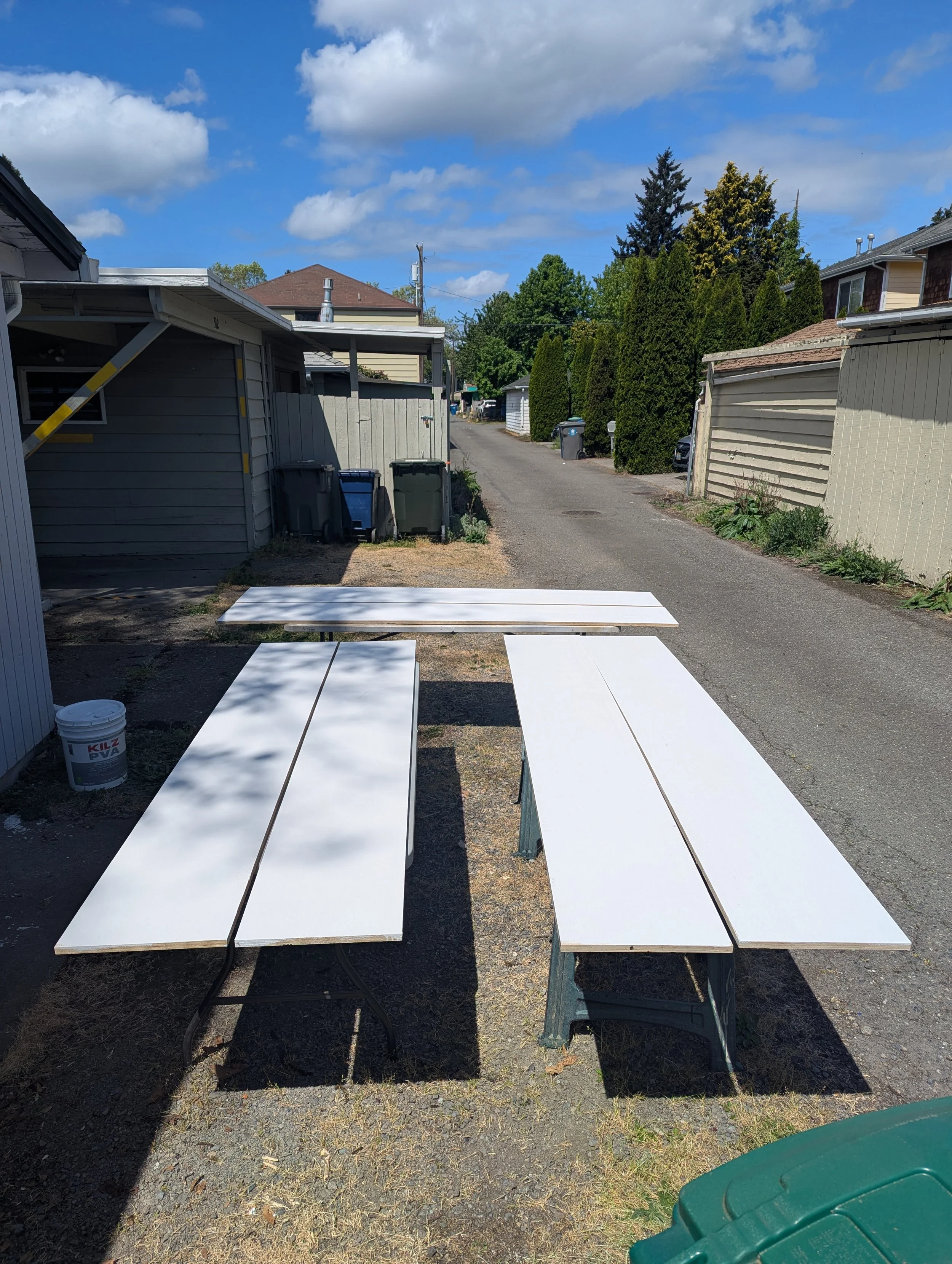 Several white-painted wooden tables and benches set up outdoors on a dirt and grass surface in a backyard, with houses, trees, and blue sky with clouds in the background.