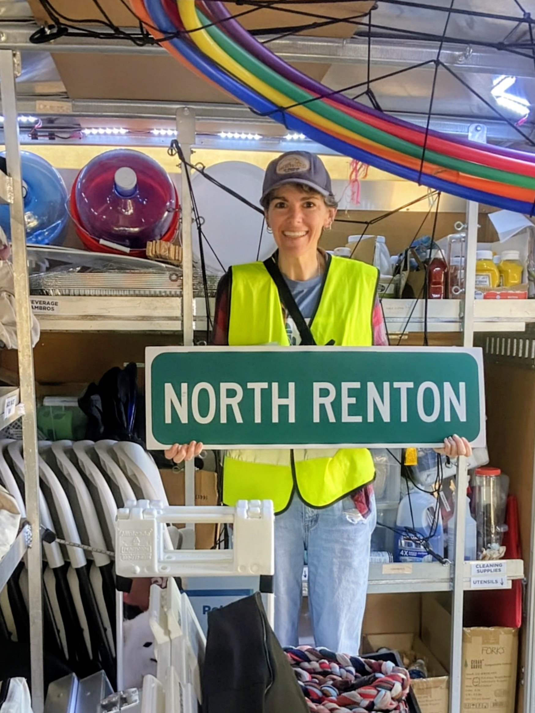 A woman wearing a baseball cap and a fluorescent safety vest is holding a North Renton sign inside a storage or retail space with shelves of cleaning supplies, plastic chairs, and various items in the background.