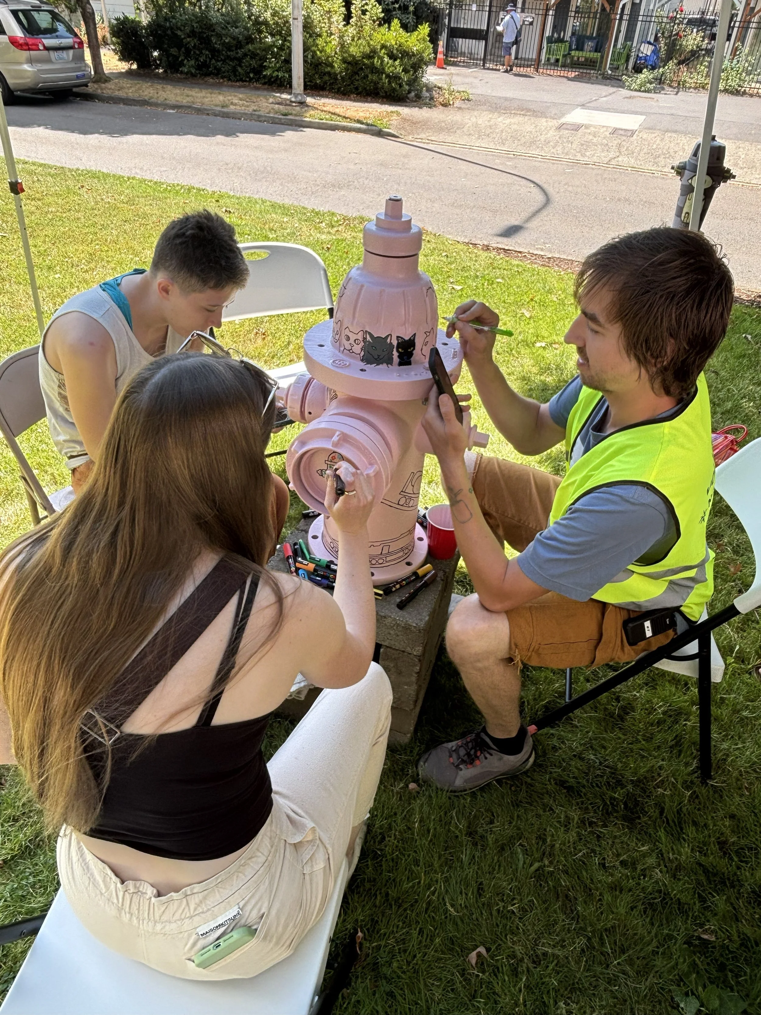 People gathered around a pink fire hydrant that is being painted with black and gray cat faces using markers, outdoors under a tent, on a grassy area.
