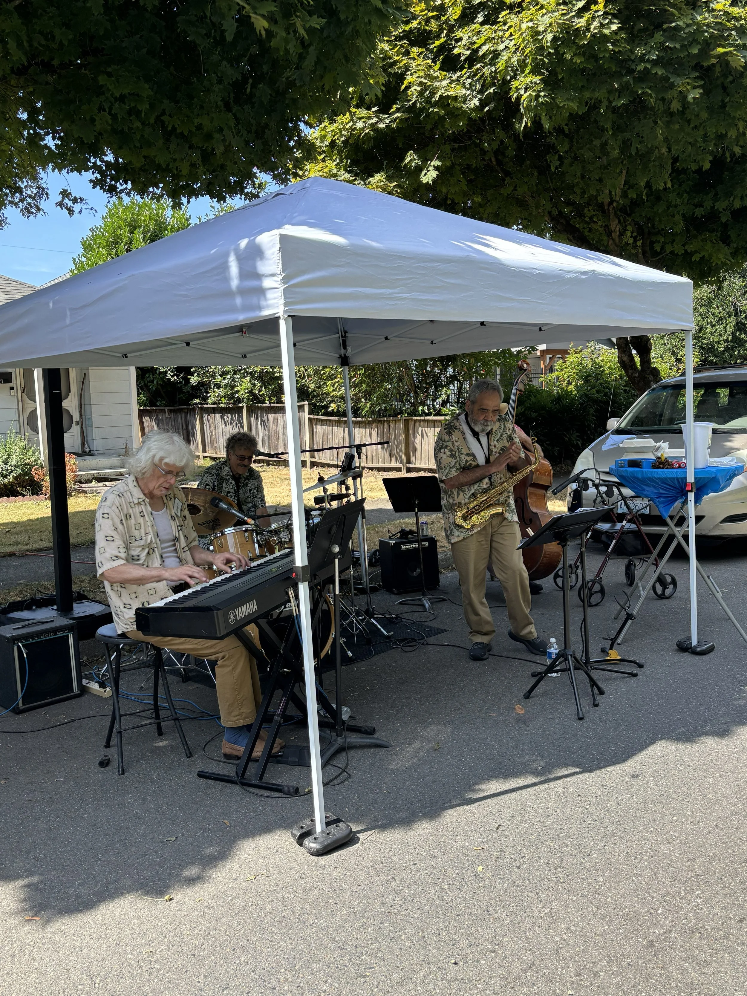 A small outdoor band performing under a white canopy on a street, with three musicians playing keyboard, drums, and saxophone. There is a white car and a wooden fence in the background, and a large tree providing shade.