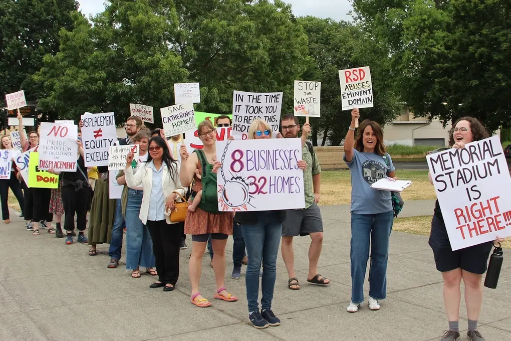 A group of people protesting outdoors, holding various signs about displacement, property rights, and local issues.