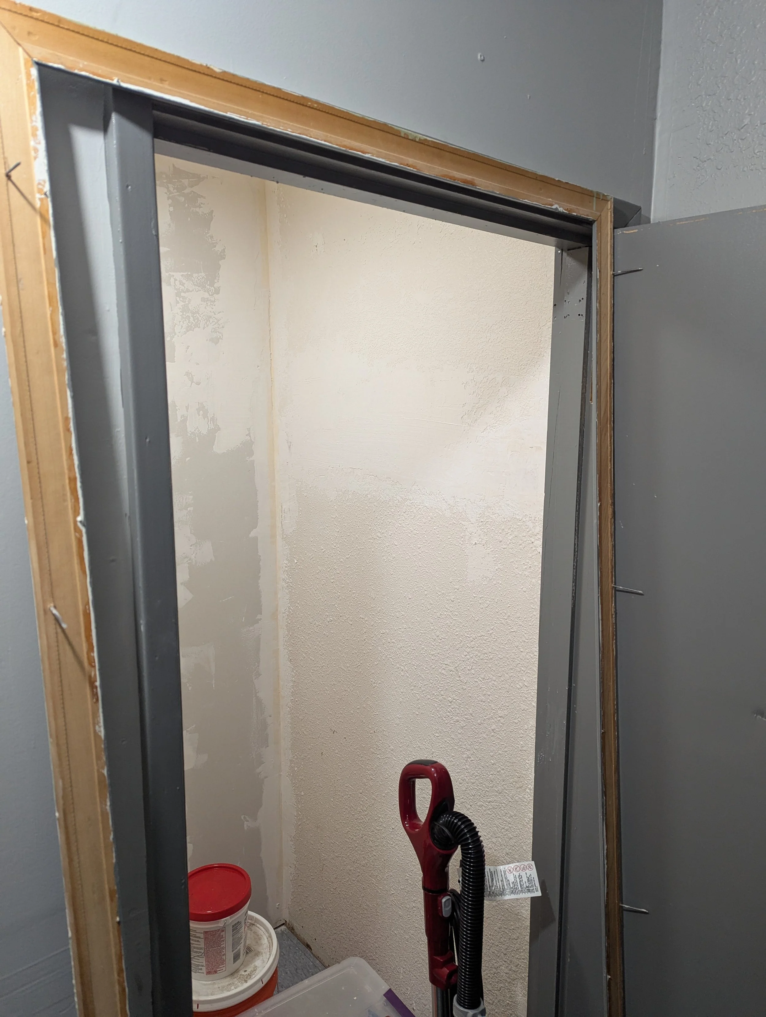View of a small unfinished closet with an open wall frame, gray metal studs, beige textured walls, and cleaning supplies including a red vacuum cleaner and two large white buckets with red lids.