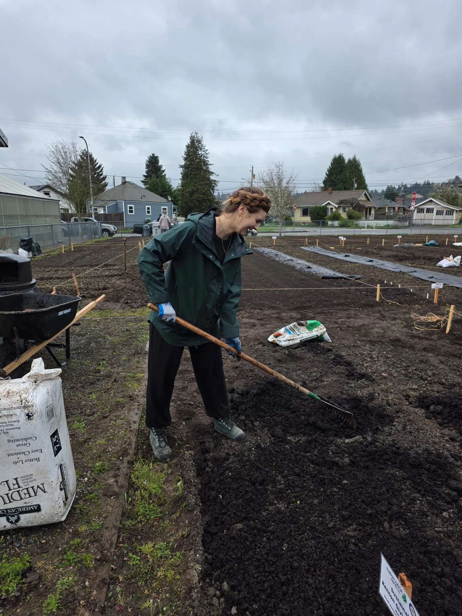 A woman with braided hair and a green jacket is working in a garden bed during a cloudy day. She is smiling and using a rake to spread soil. There are houses and trees in the background, along with gardening supplies and a sign.