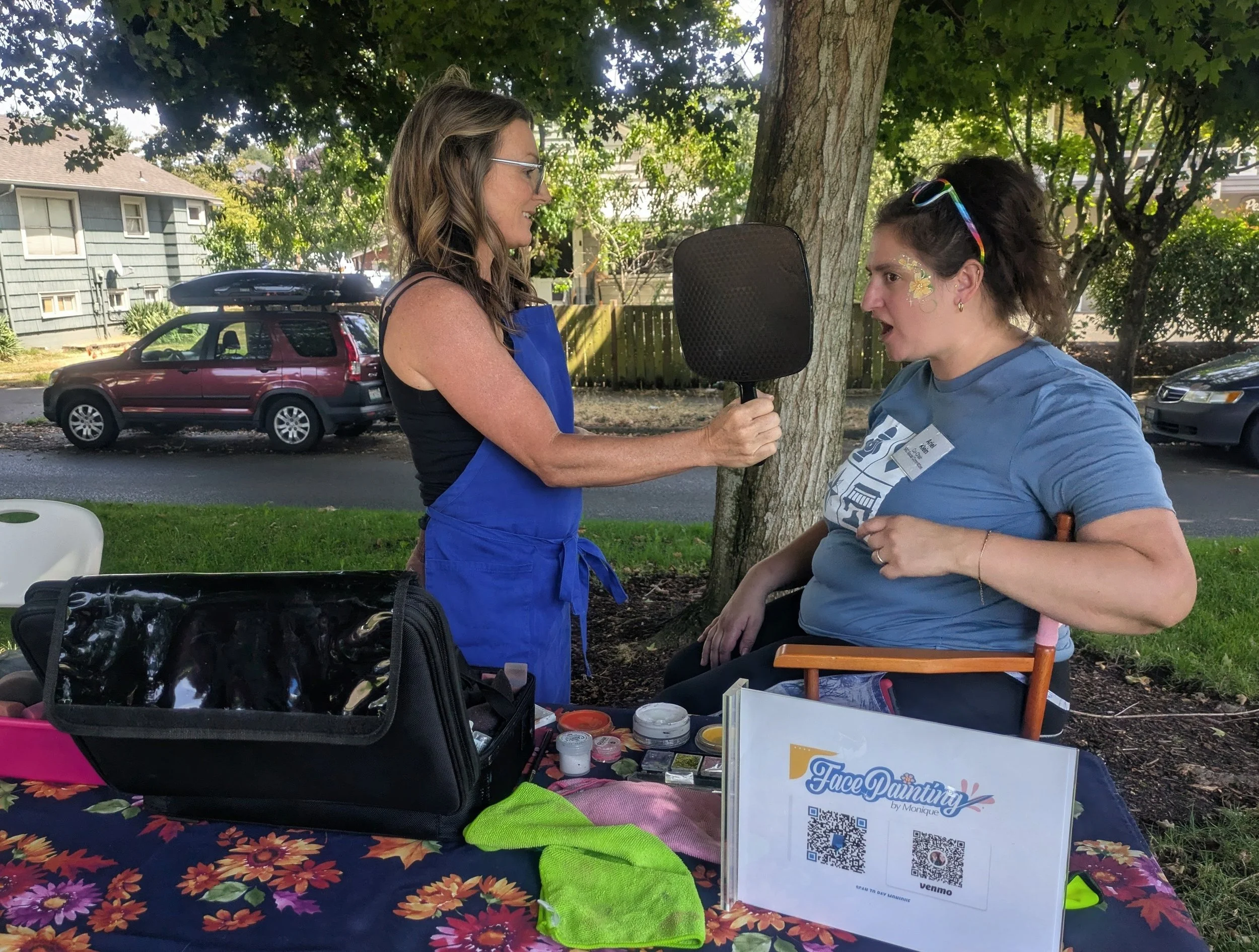 Two women at an outdoor face painting booth. One woman with glasses and long wavy hair interviews or talks to the other woman, who has earrings, a floral face painting, and sunglasses on her head. The booth has a table with supplies like paint, brush