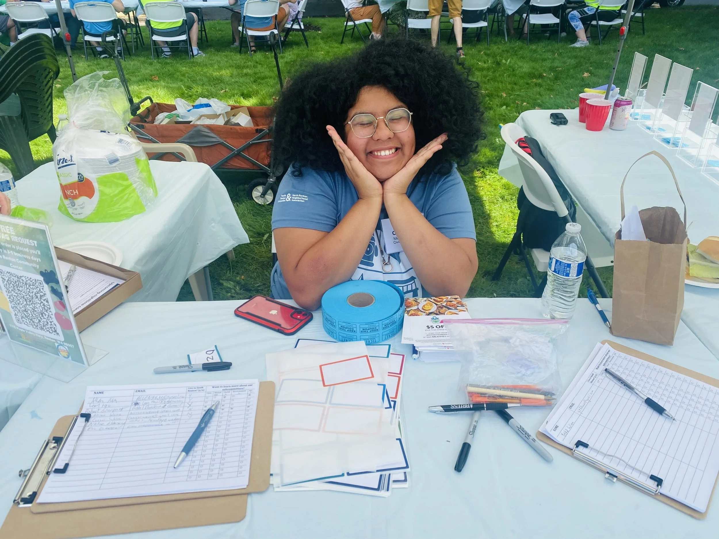 A woman with glasses and curly hair smiling at a table outdoors, surrounded by papers, pens, a water bottle, and other items, with a group of people sitting at tables in the background.