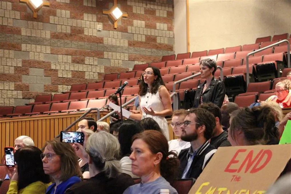A woman standing and speaking at a microphone during a conference or public event, with attendees seated and some recording with smartphones in an auditorium with brick walls.