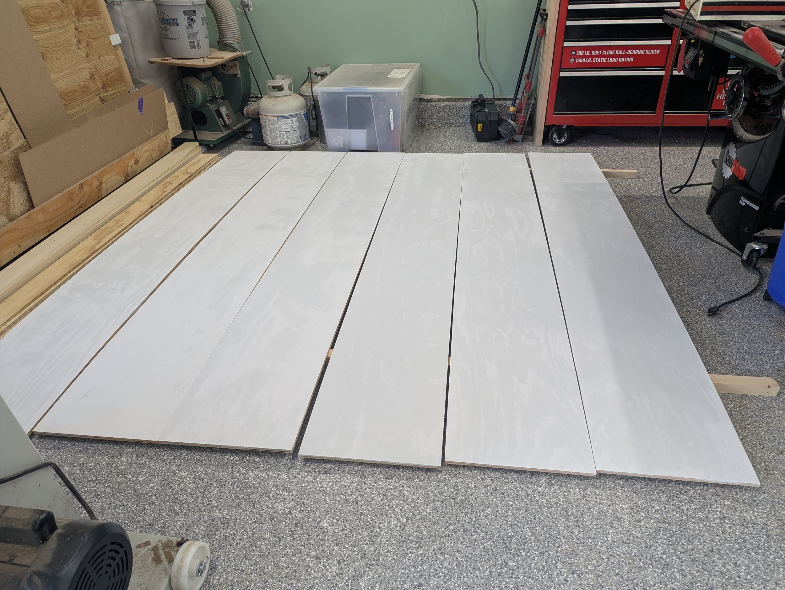Garage workspace with white painted wooden boards laid on the floor, with tools and storage containers around, including a red tool chest and a propane tank.