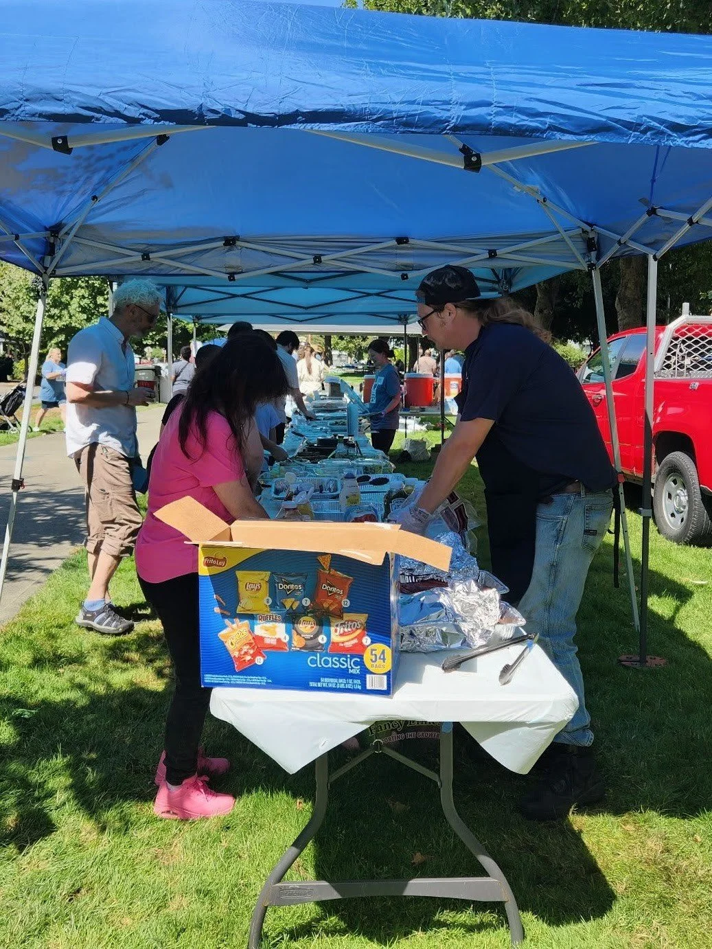 People at an outdoor food stall with a blue canopy, serving or buying food during a sunny day.