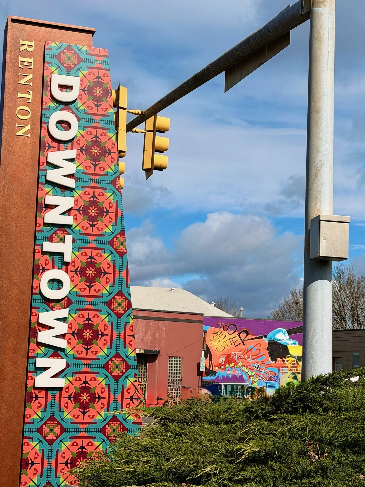 A decorative sign for Wyoming with colorful geometric patterns and large white letters spelling 'WYOMING'. Behind the sign, there are colorful protesters banners with graffiti art, a building with a metal roof, and a blue sky with some clouds.