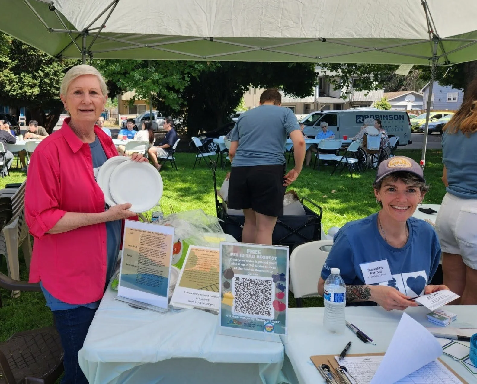 Two women sitting at a table under a canopy at an outdoor event. The woman on the left, with short blonde hair, wearing a pink shirt, is holding white plates and standing next to informational flyers on the table. The woman on the right, with short g