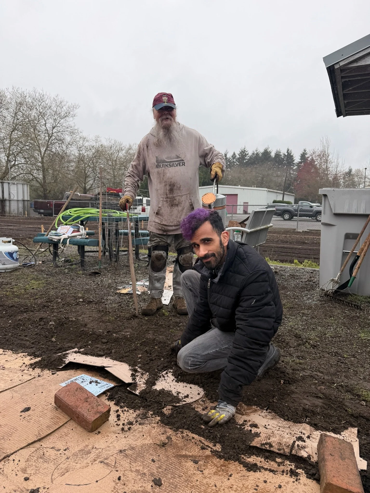 Two men working in a garden or farm plot, with one kneeling and the other standing and holding gardening tools. The area is prepared for planting, with soil, paving, and some bricks visible. The background shows trees, buildings, and parked vehicles 
