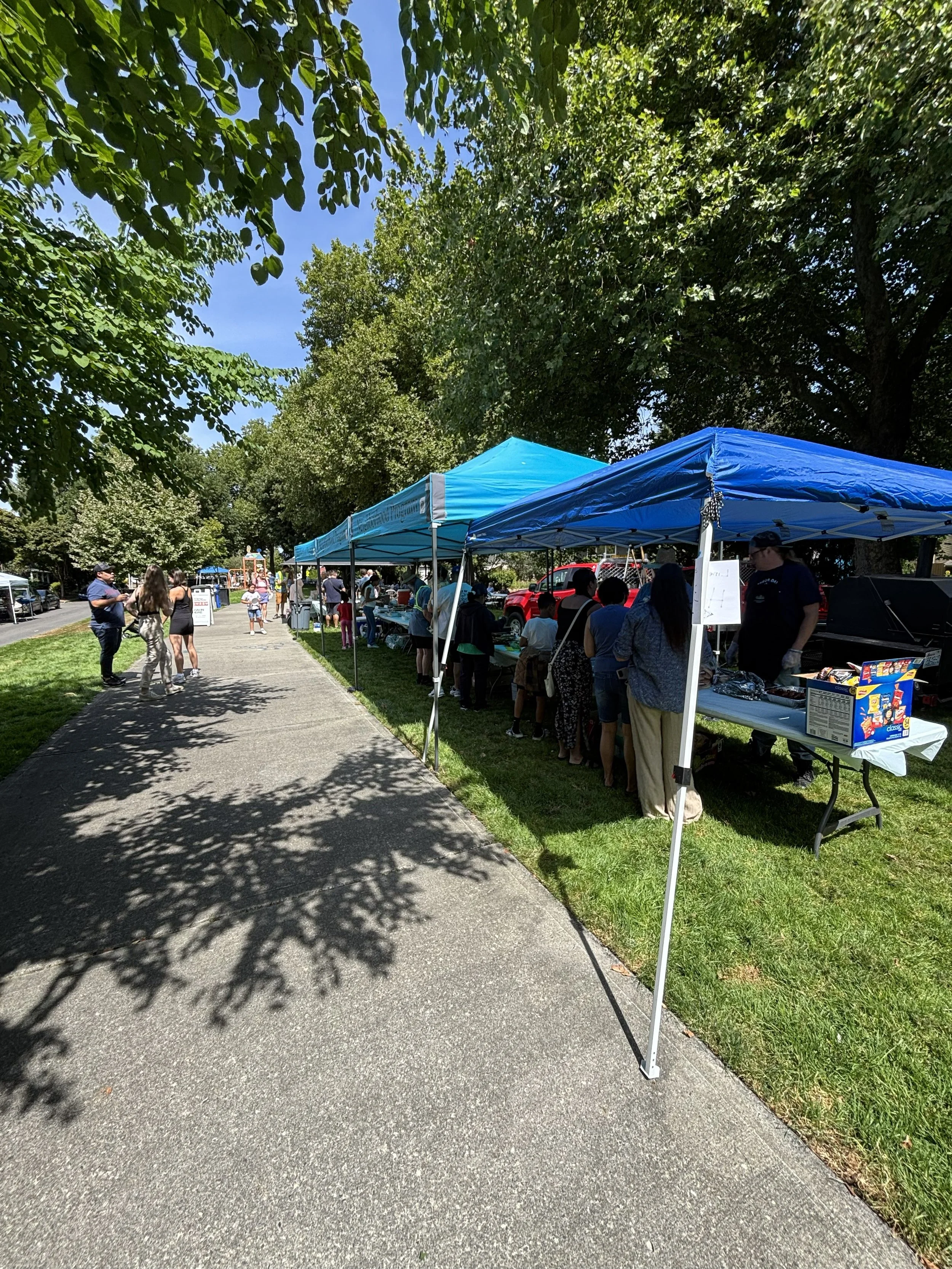 Community event in a park with blue tents, people shopping and socializing, trees casting shadows on the sidewalk.