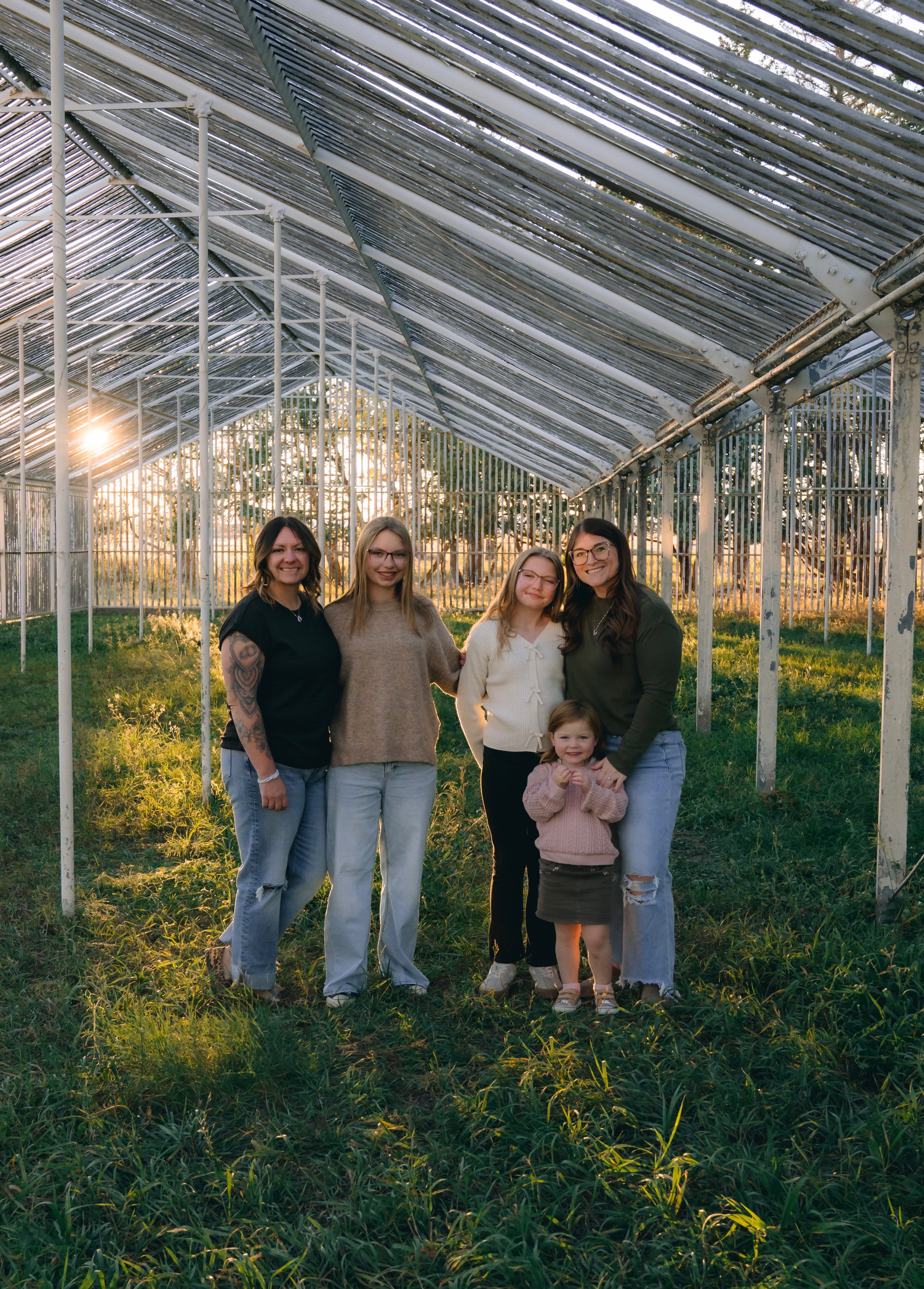 Five women and a young girl standing inside a greenhouse during sunset, smiling and posing for the photo.