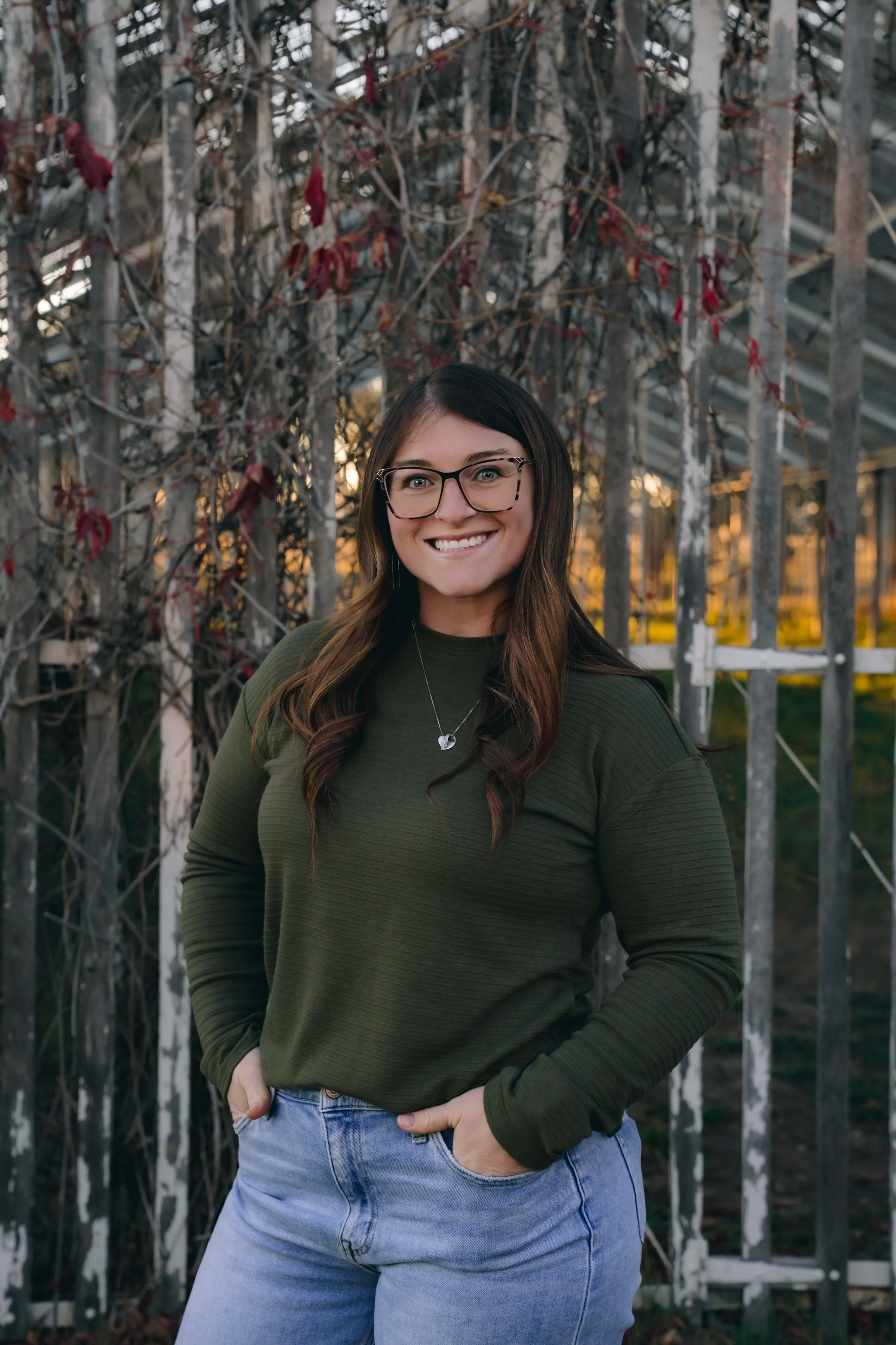 A smiling woman with glasses wearing an olive green sweater and light blue jeans, standing outdoors in front of a metal fence and autumnal vine with red leaves.