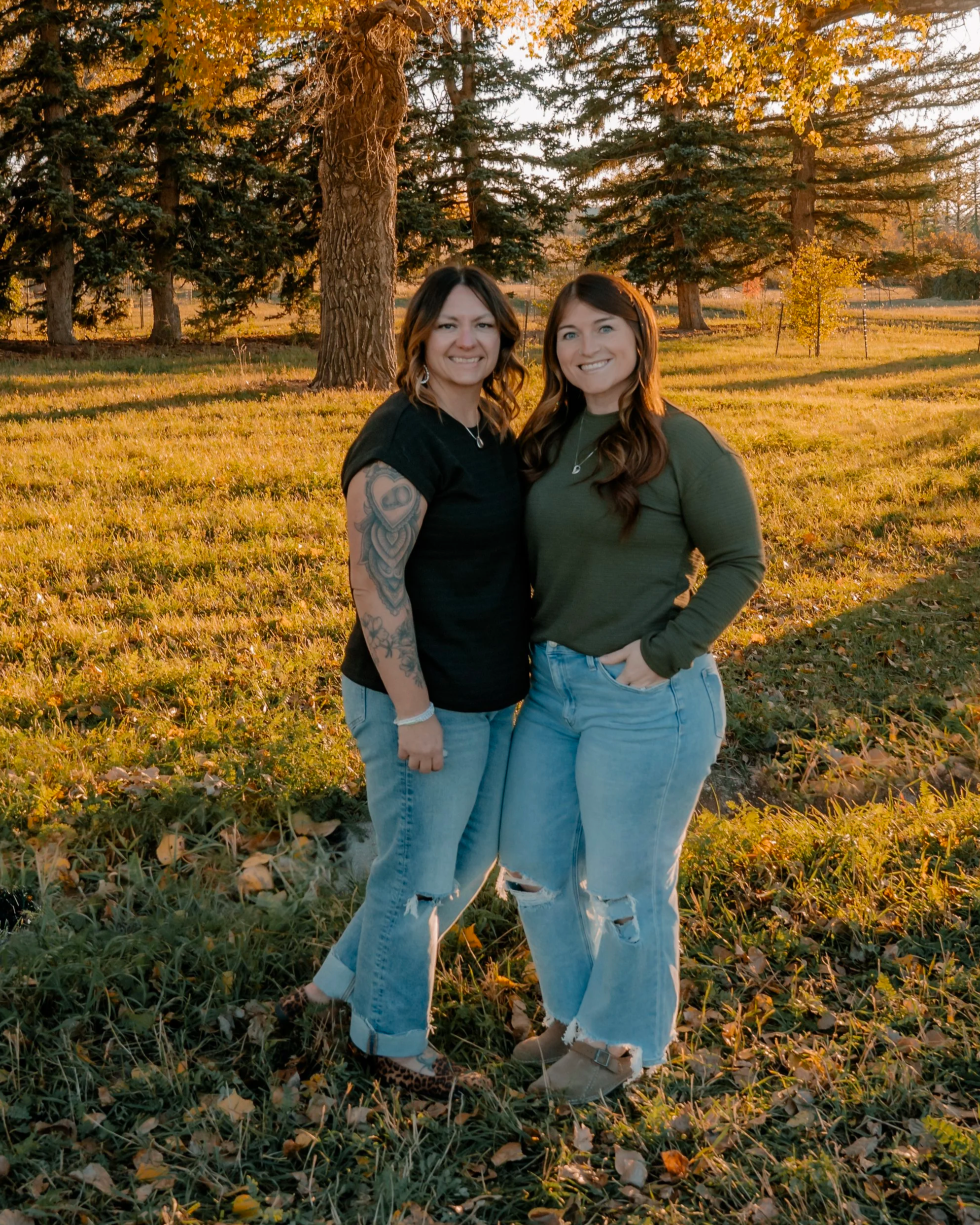Two women standing together outdoors in a grassy park during sunset, with trees in the background. One woman has shoulder-length brown hair, tattoos on her right arm, and is wearing a black shirt and ripped jeans. The other has long brown hair, and is wearing a green sweater and ripped jeans. Both are smiling.