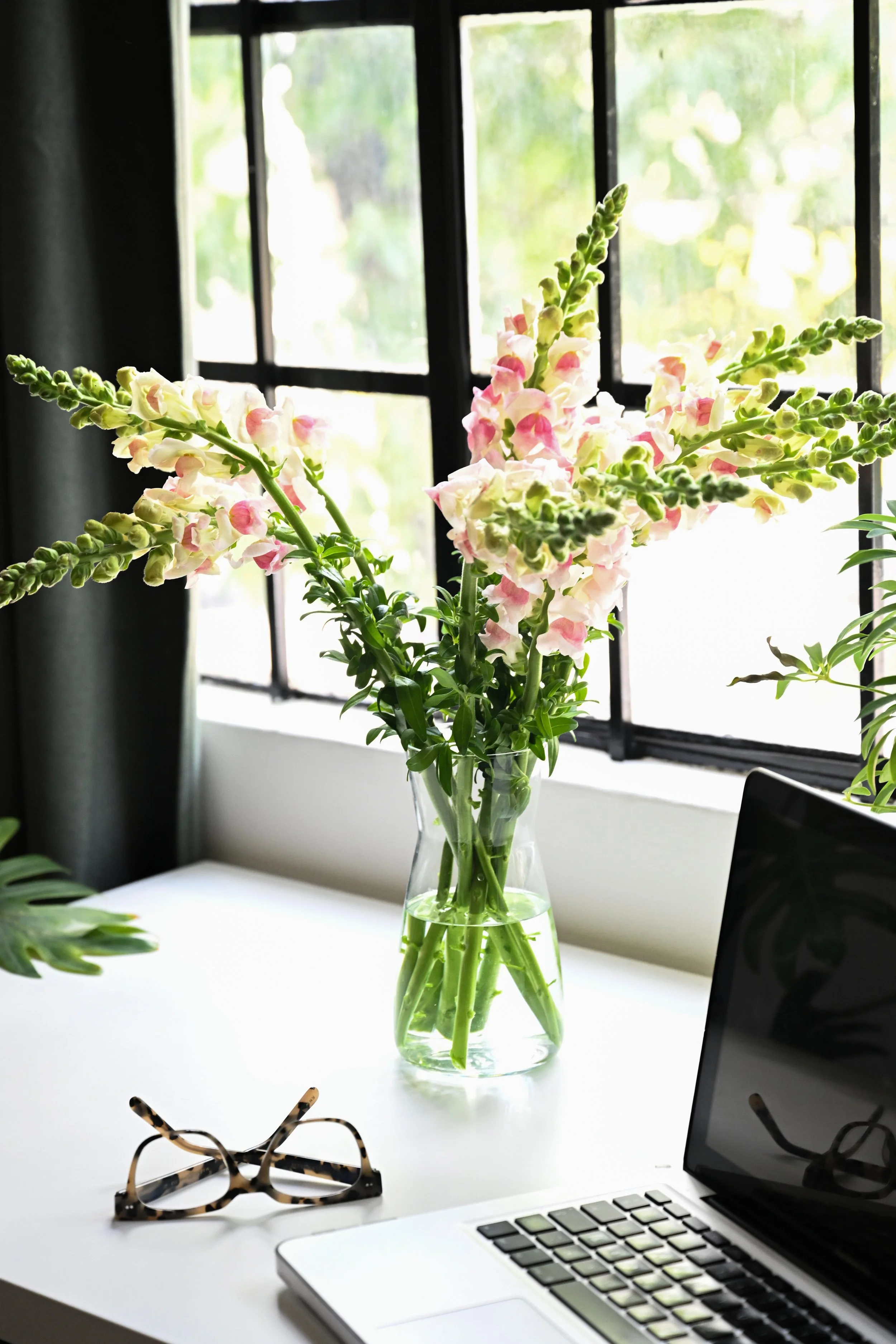 Een venster met een vaas met bloemen, een laptop en een bril op een witte tafel.