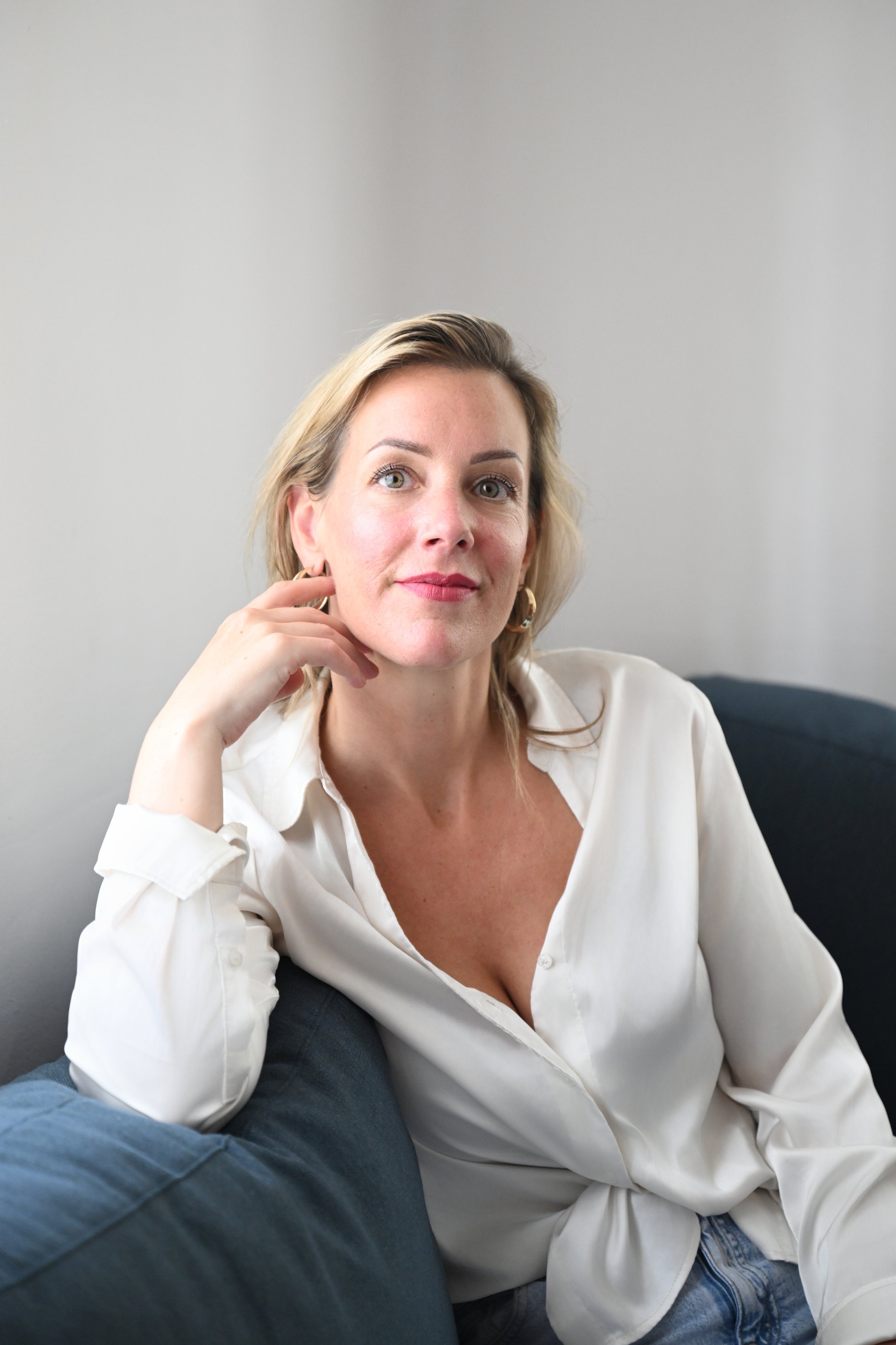 Portret van een vrouw met blonde haar die op een bank zit, witte blouse, lichte achtergrond.