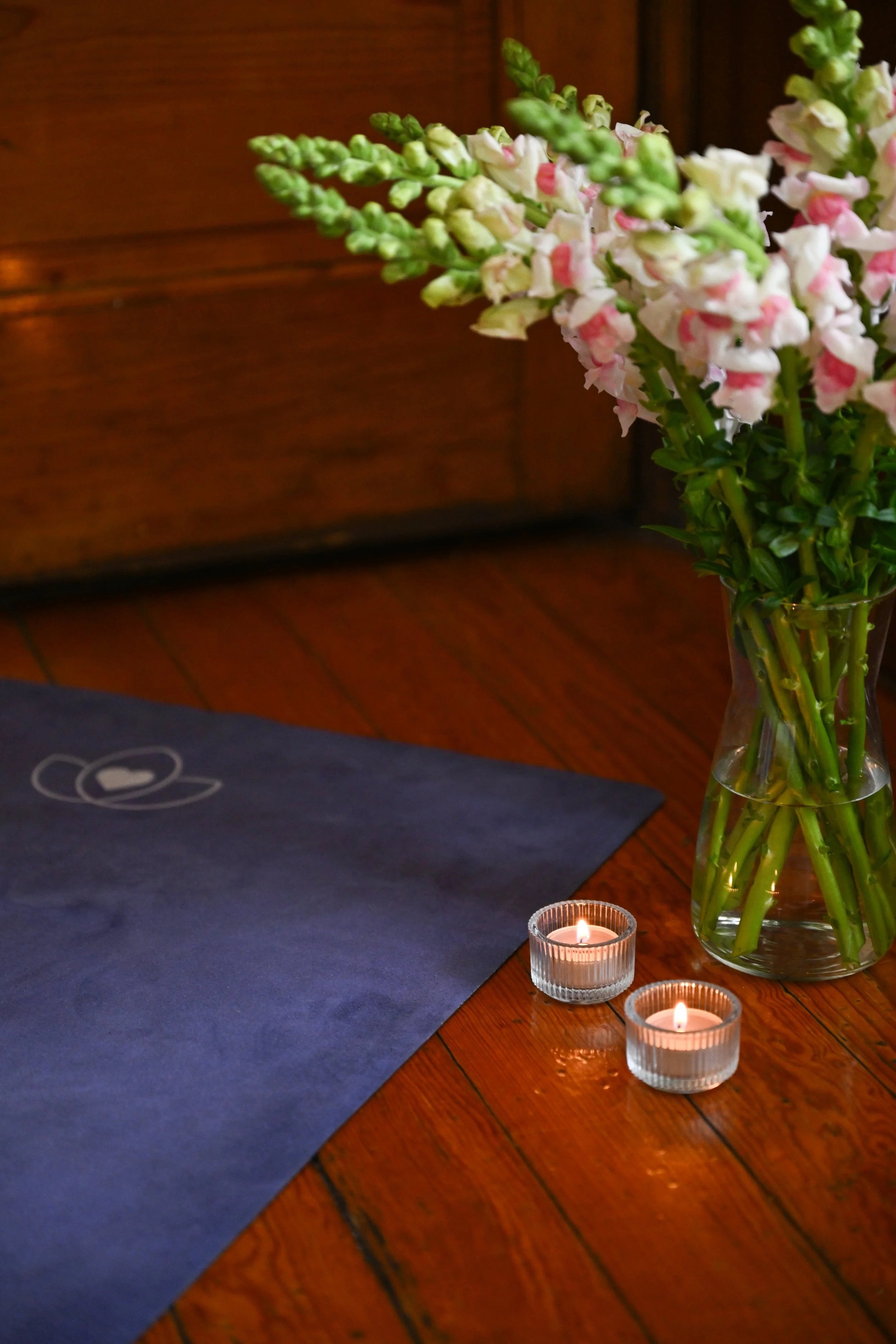 Vaas met gepinkte bloemen, twee kleine brandende kaarsen op een houten vloer, en een donkerblauwe placemat met een witte hart- en cirkeltekening.