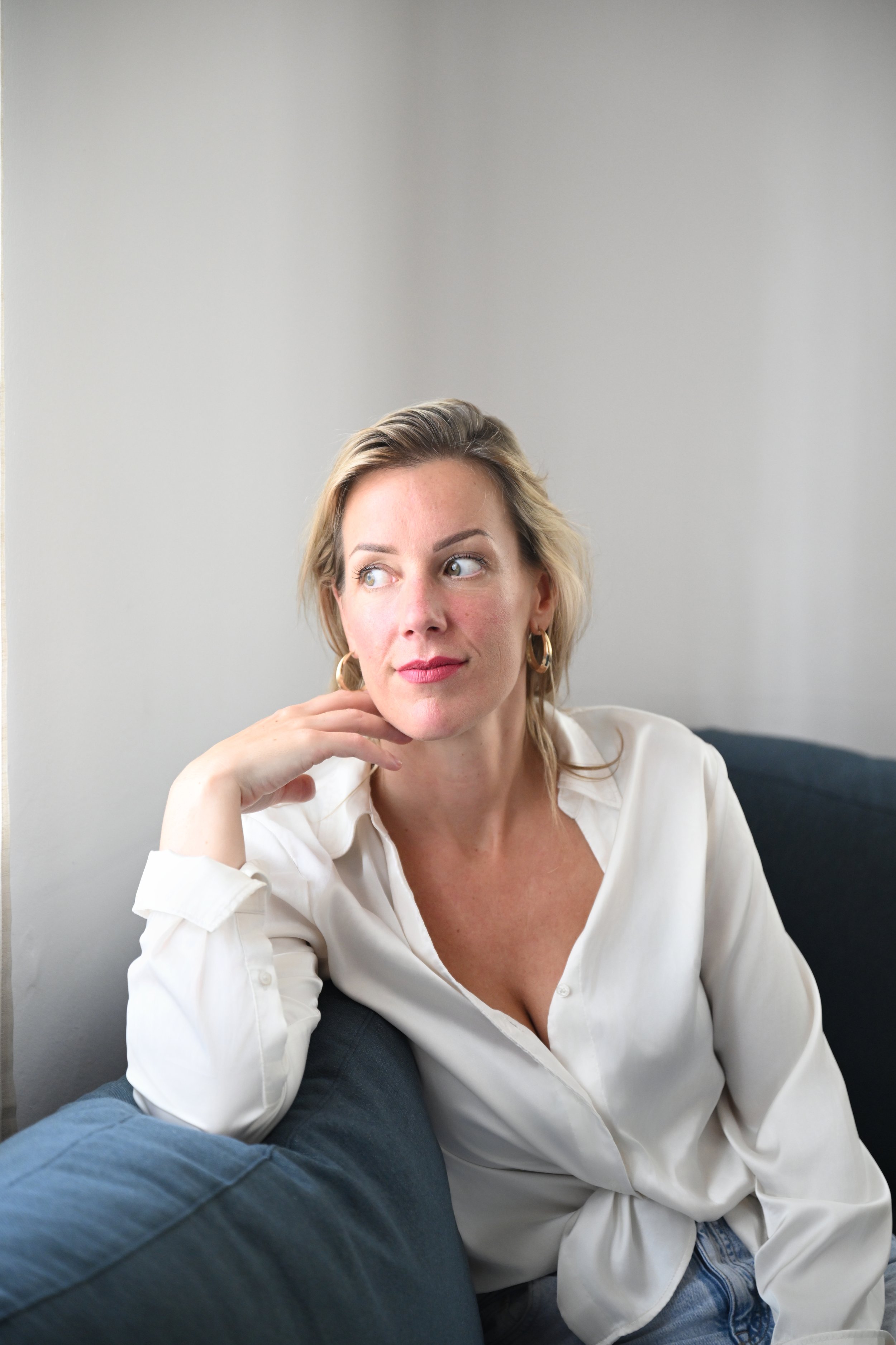 Portret van een vrouw met blond haar die op een bank zit, draagt een witte blouse en kijkt naar links.