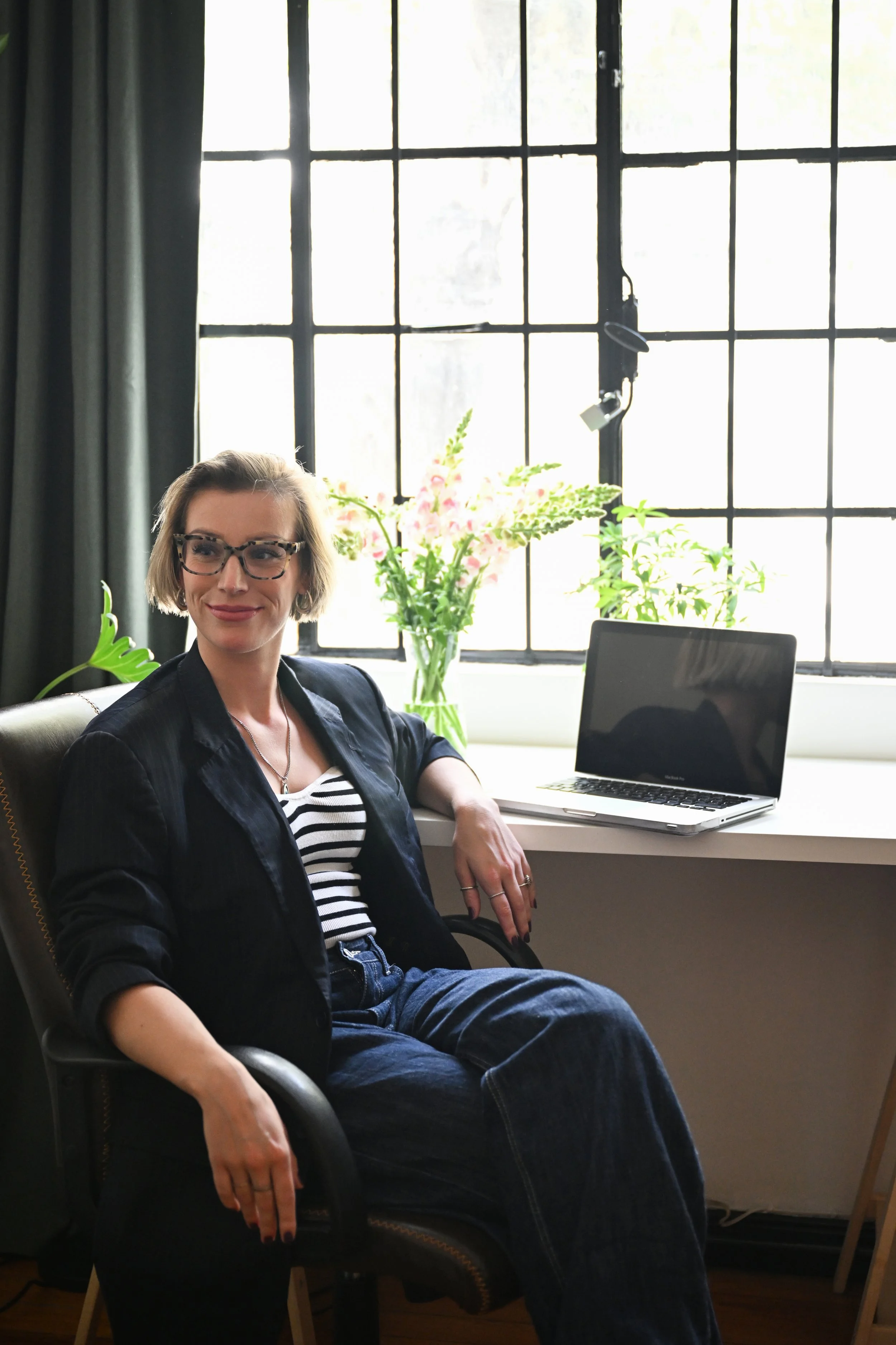 Een vrouw met kort bruin haar en glazen, zittend in een zwarte bureaustoel naast een bureau met een open laptop en een vaas met bloemen, voor een raam met zwart kozijn en groene planten.