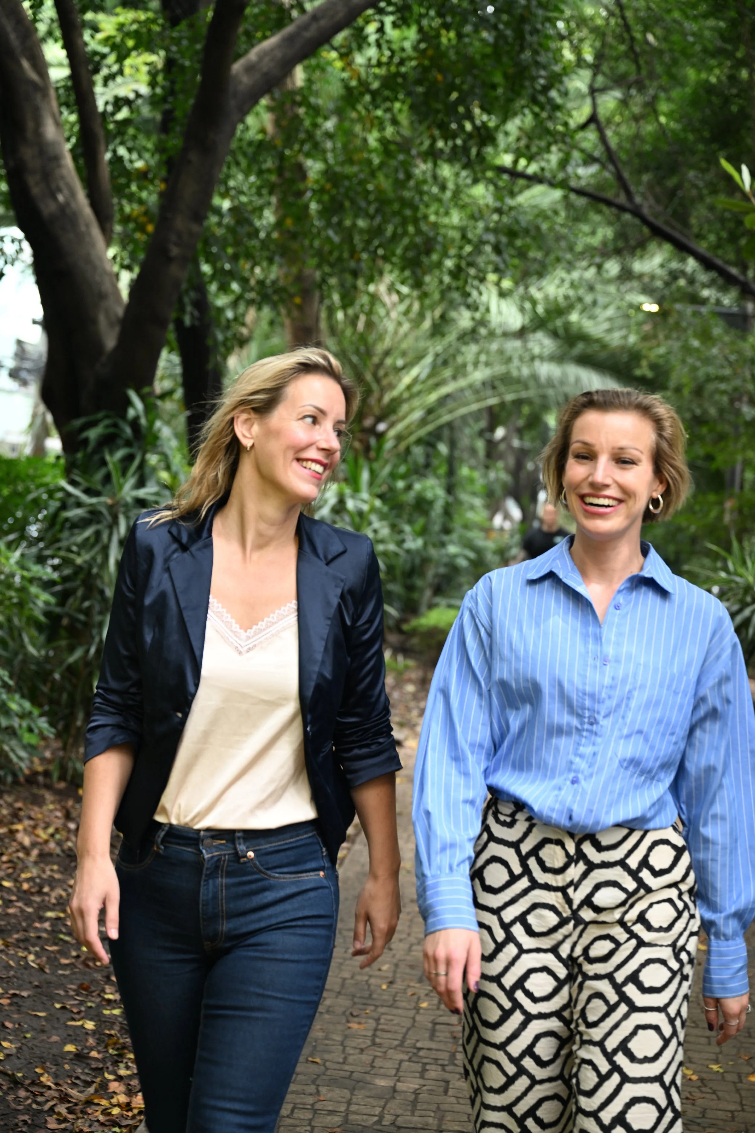 Twee vrouwen lopen lachend door een groene tuin of park, omgeven door bomen en struiken, op een tegelpad.
