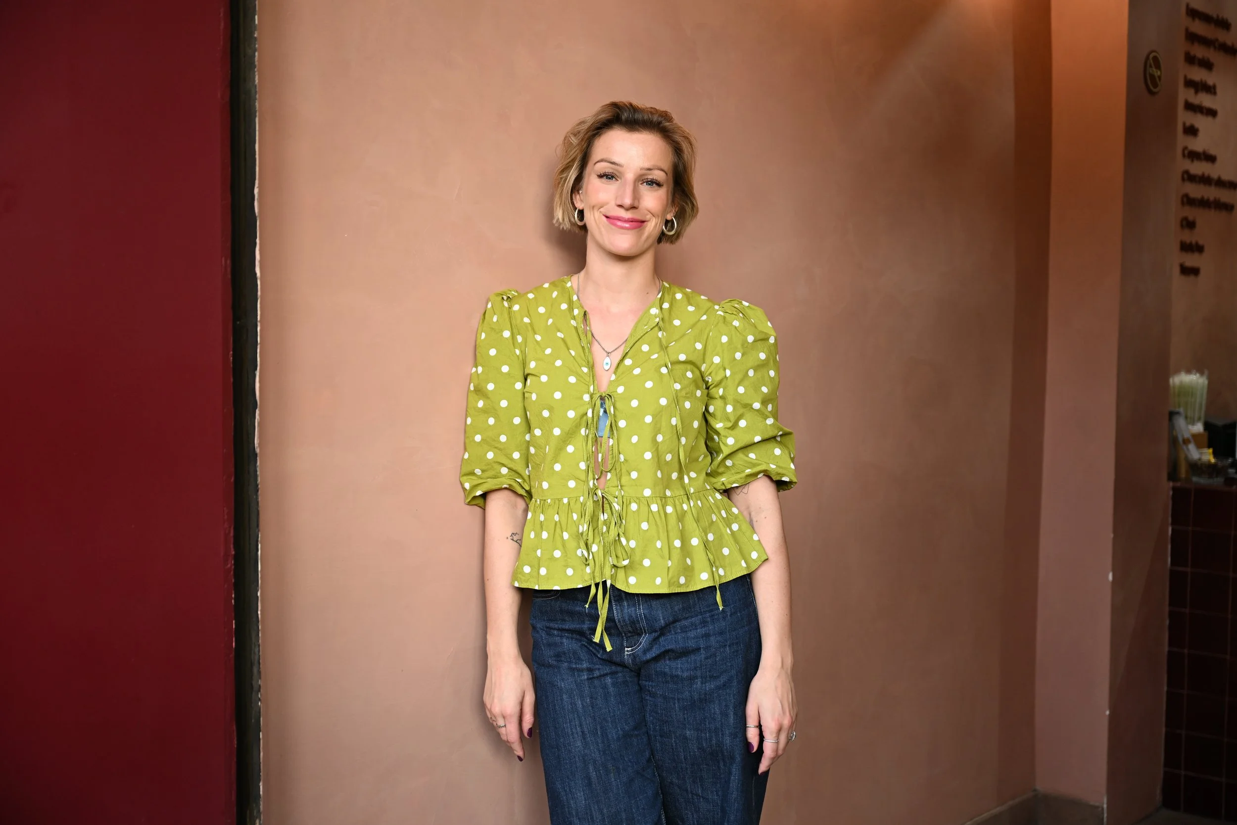Vrouw met blonder haar, lachend, in een groene blouse met witte stippen en blauwe jeans, staand voor een beige muur.