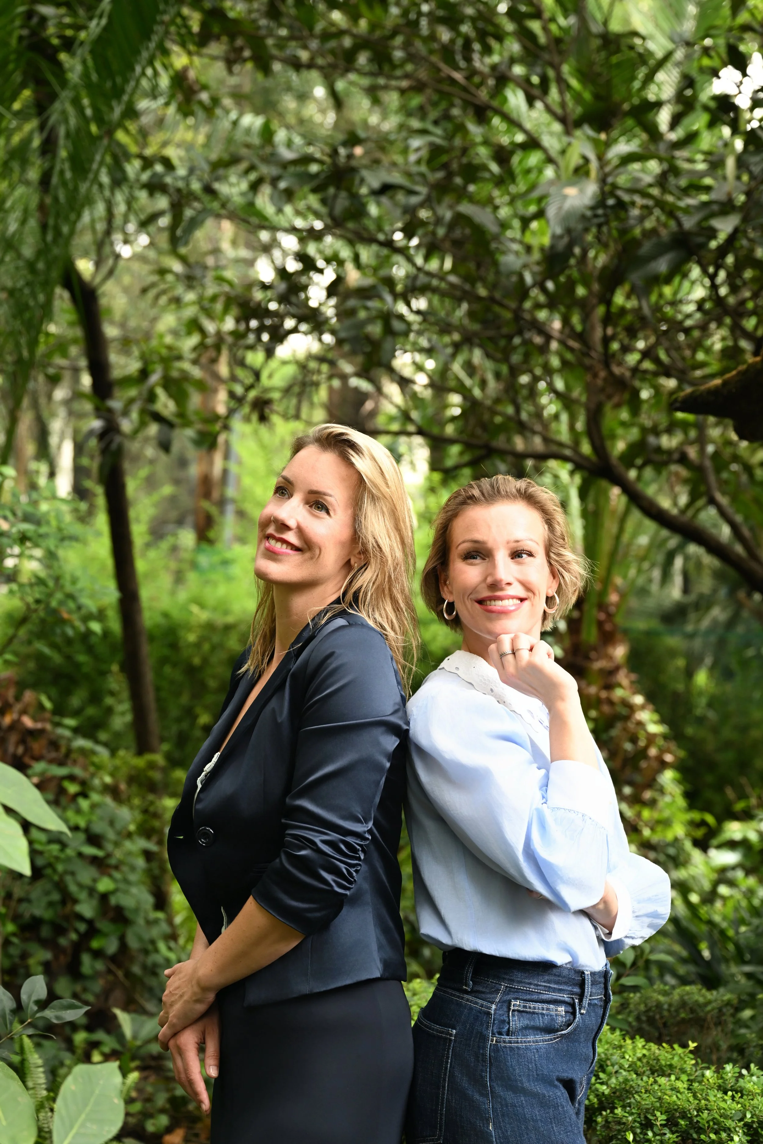 Twee vrouwen staan in een groene tuin, lachend en leunend tegen elkaar.